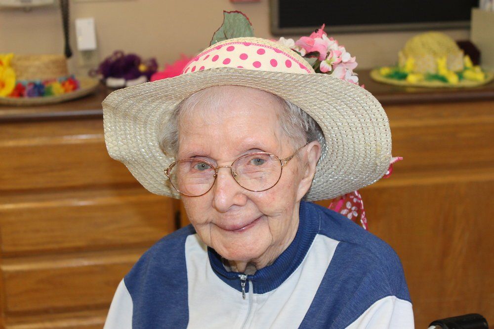 Elderly woman smiles, wearing a straw hat with pink polka dots and flowers; indoor setting.
