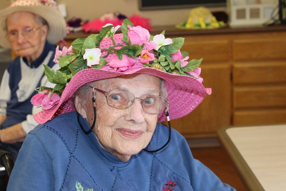 Elderly woman in blue sweater and floral pink hat smiles.