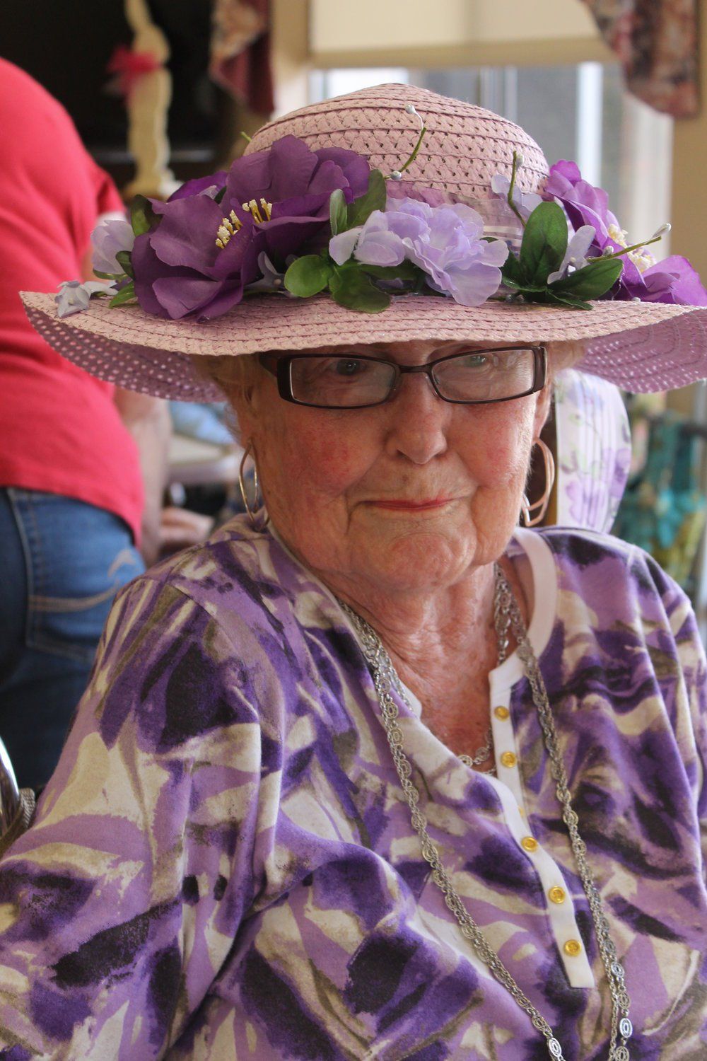 Elderly woman in purple floral hat and sweater, wearing glasses, smiling.