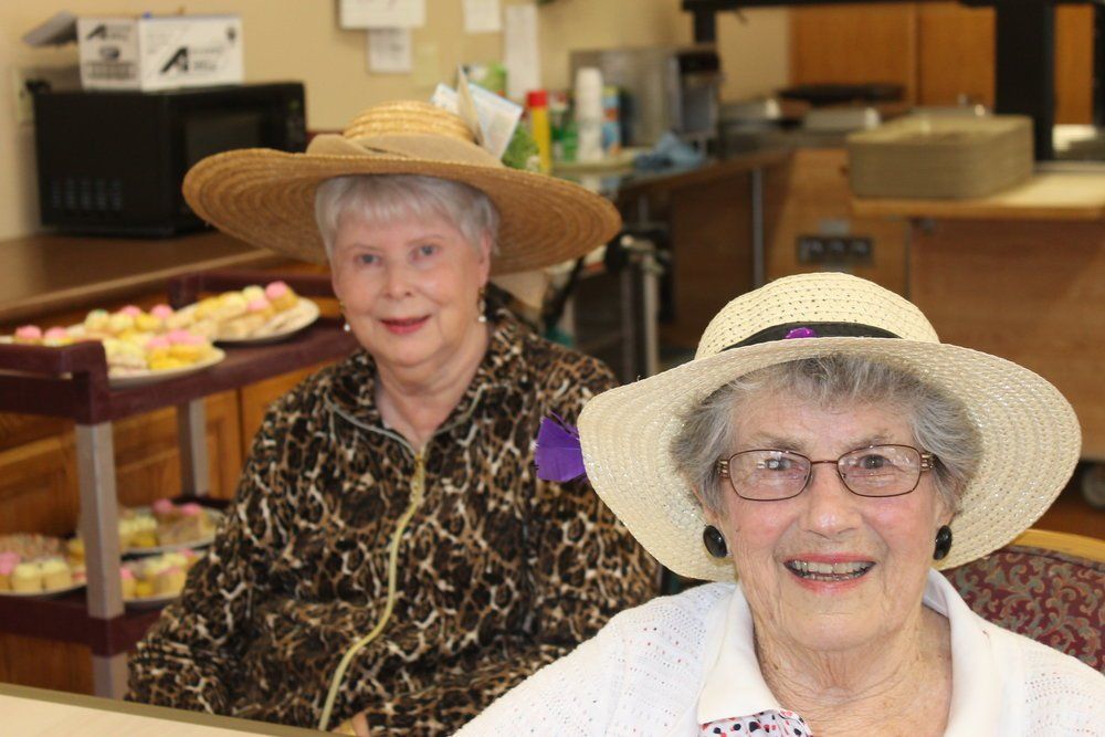 Two elderly women in hats smile at the camera; a table of treats is in the background.