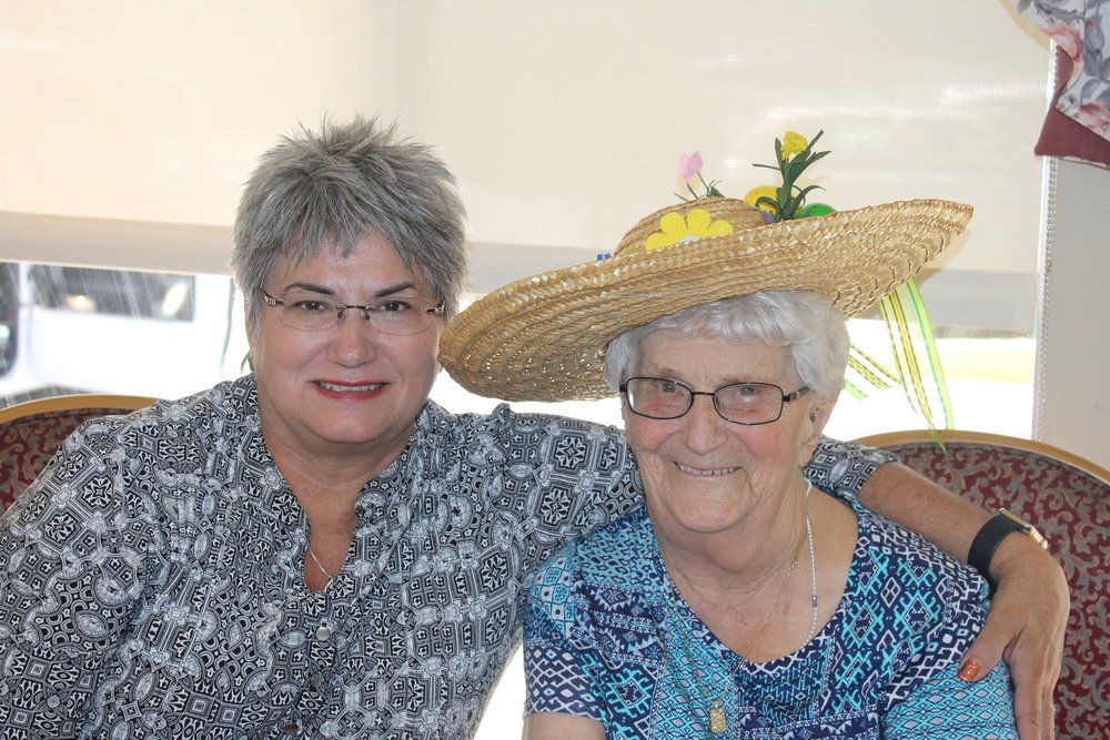 Two women smiling, one wearing a straw hat with floral decorations, indoors.