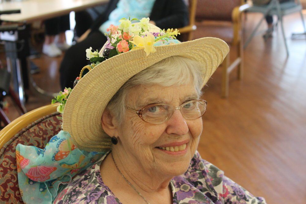 Smiling elderly woman wearing a straw hat decorated with flowers; indoors, seated.