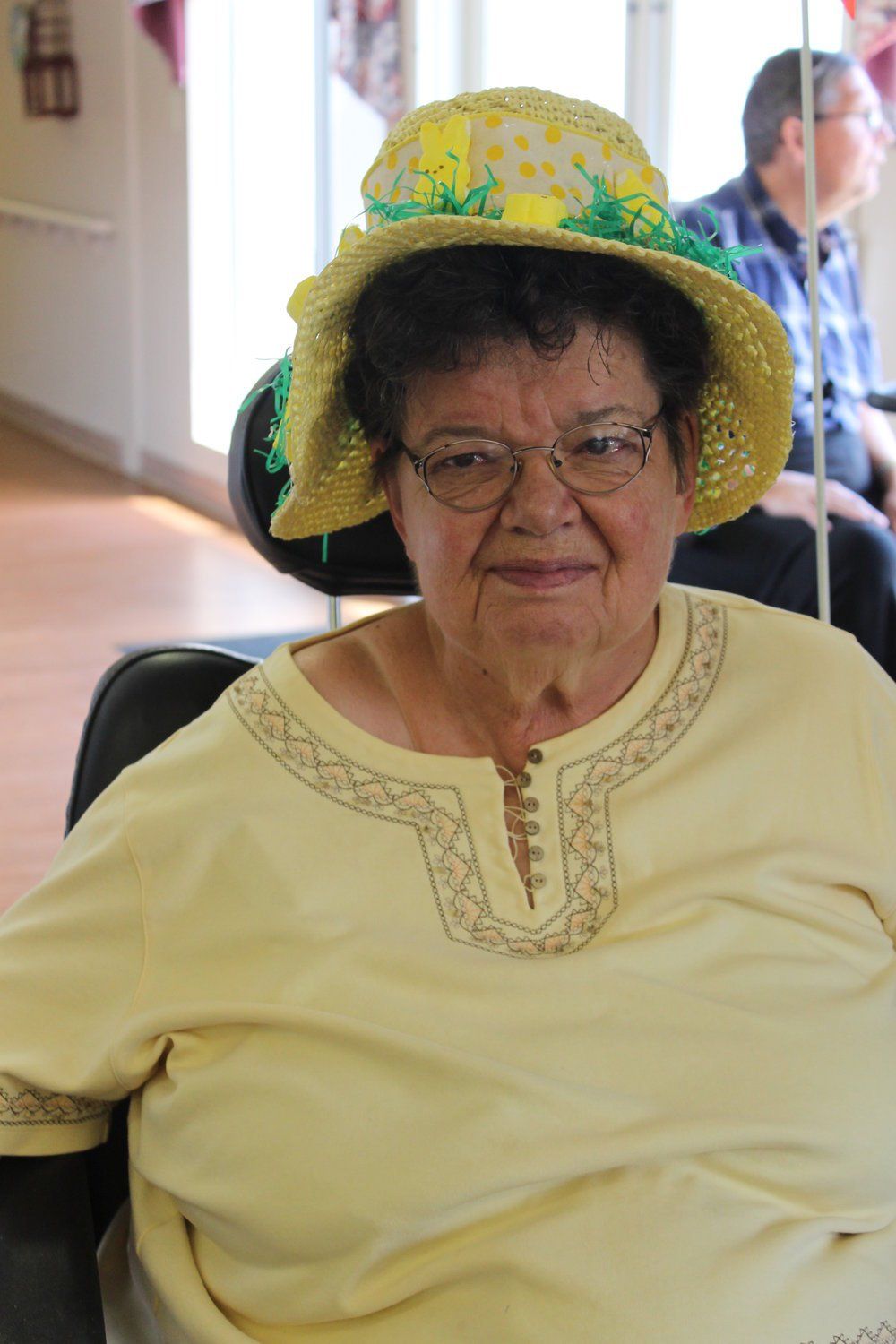 Elderly woman wearing glasses and yellow straw hat, smiling, sitting indoors.