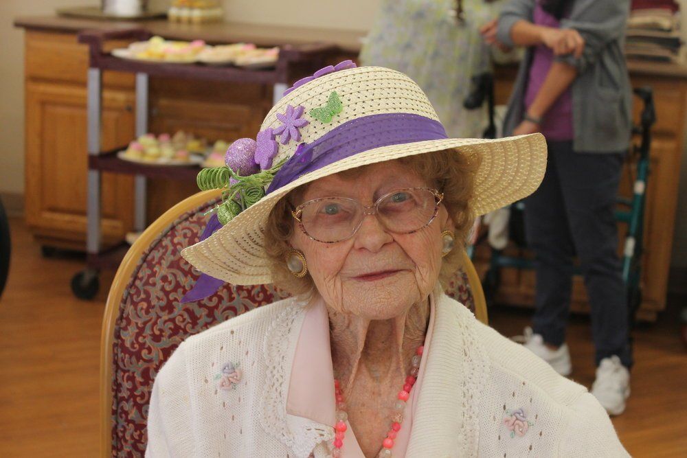 Elderly woman in a decorated hat, sitting indoors, wearing a white sweater.