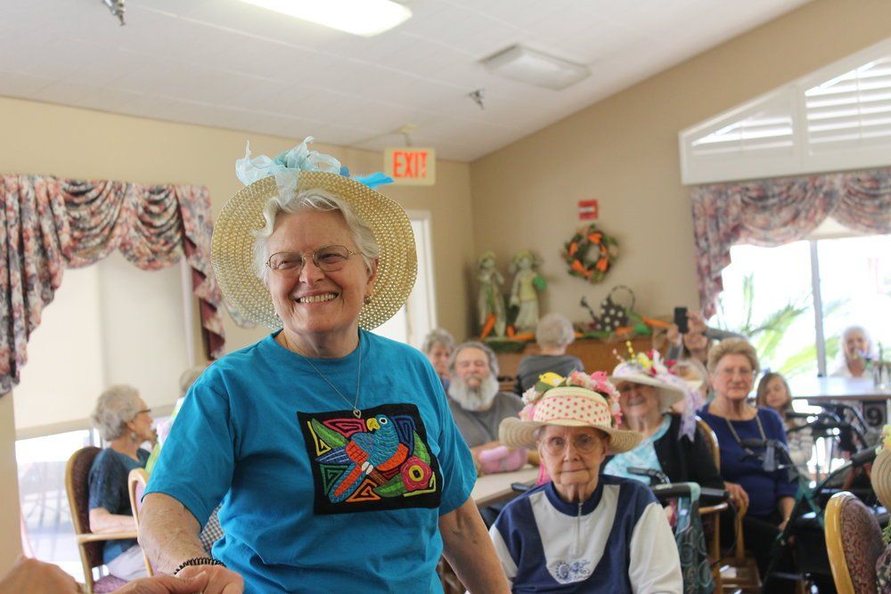 Woman in a straw hat and blue shirt smiles at a gathering of elderly people in hats.