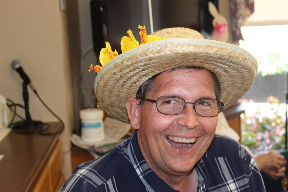 Man wearing straw hat decorated with yellow flowers, smiling and laughing.