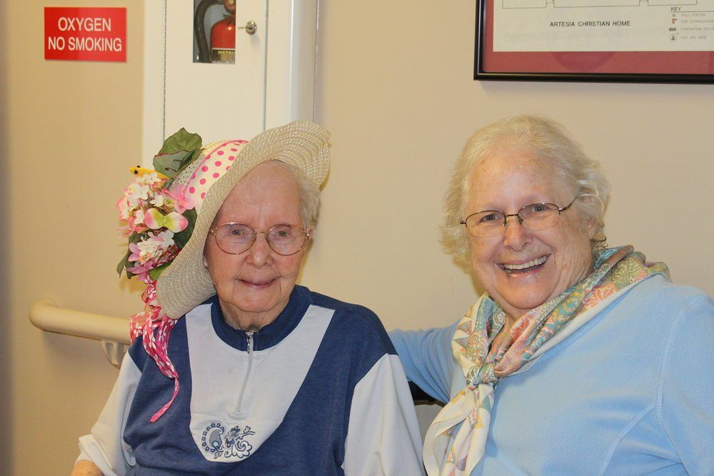 Two older women, one wearing a decorated hat, smiling indoors.