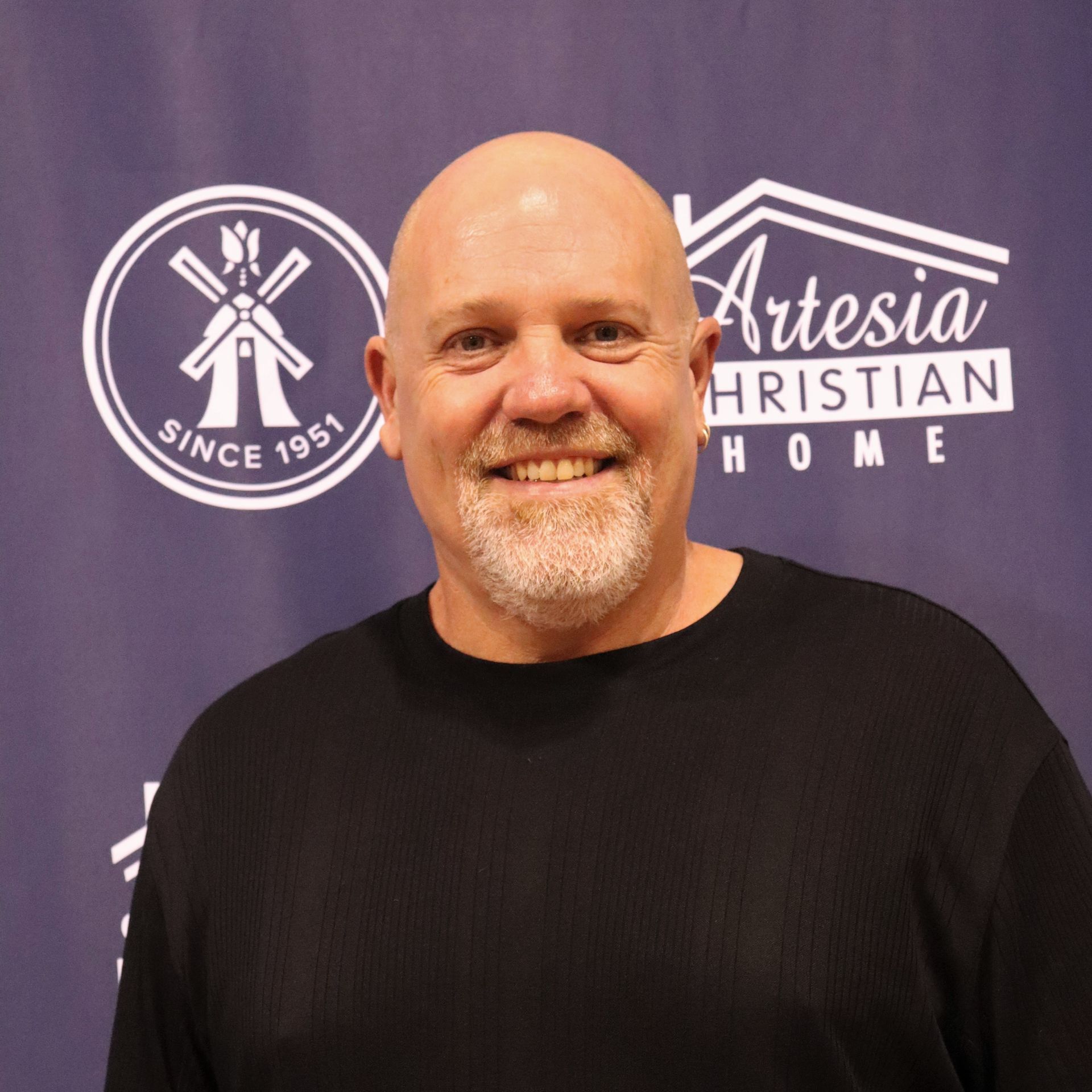 Bald man with a graying beard smiles, in front of a backdrop with logos for a Christian Home.