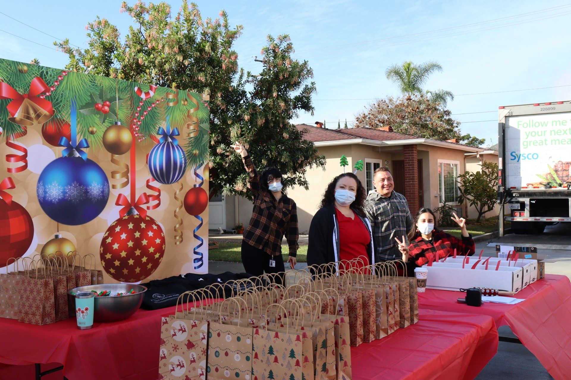 People wearing masks at an outdoor holiday event with gifts, decorations, and a truck in a residential area.