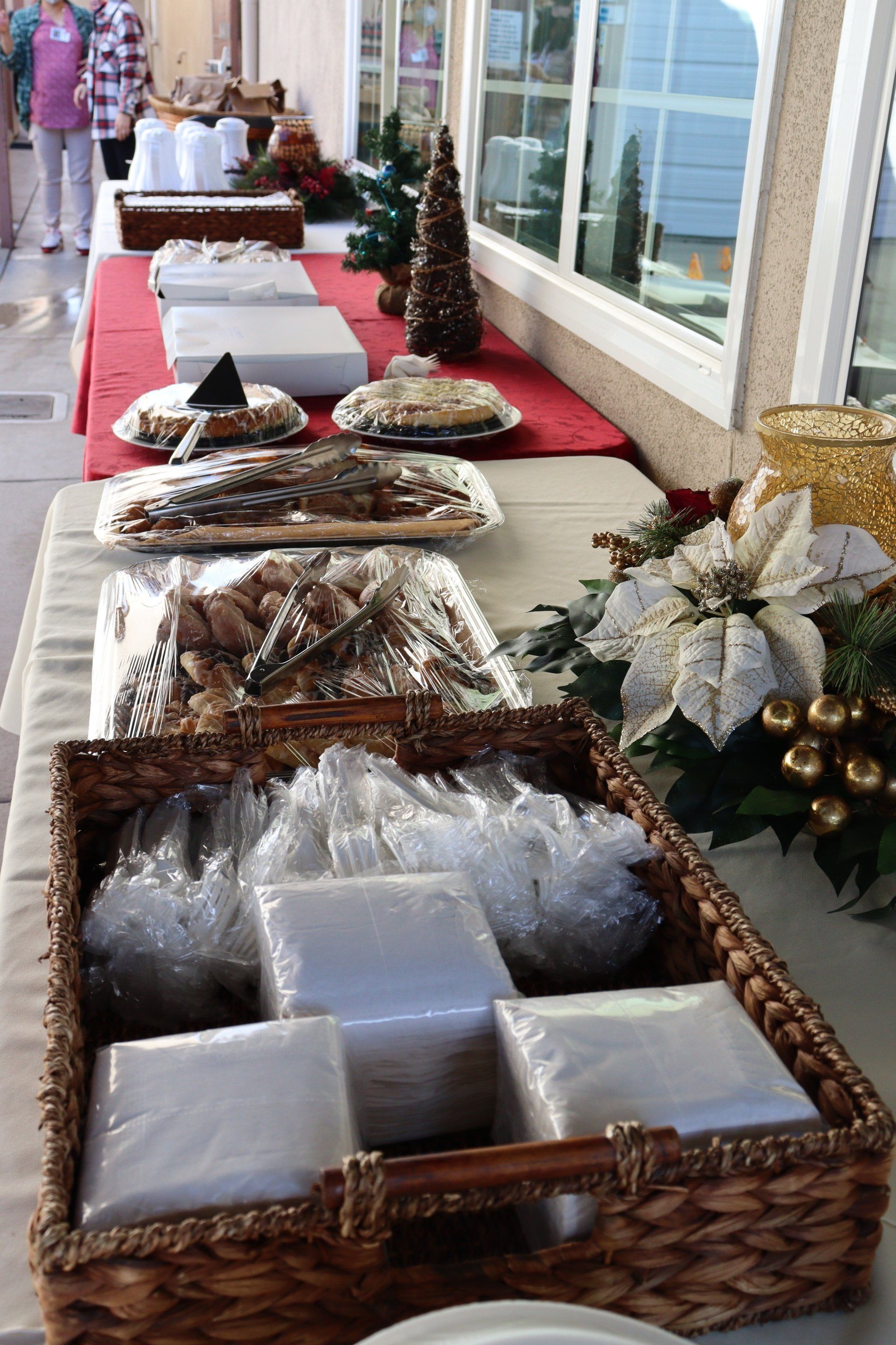 Table of food covered in plastic wrap, plus napkins and utensils, set for a holiday event.