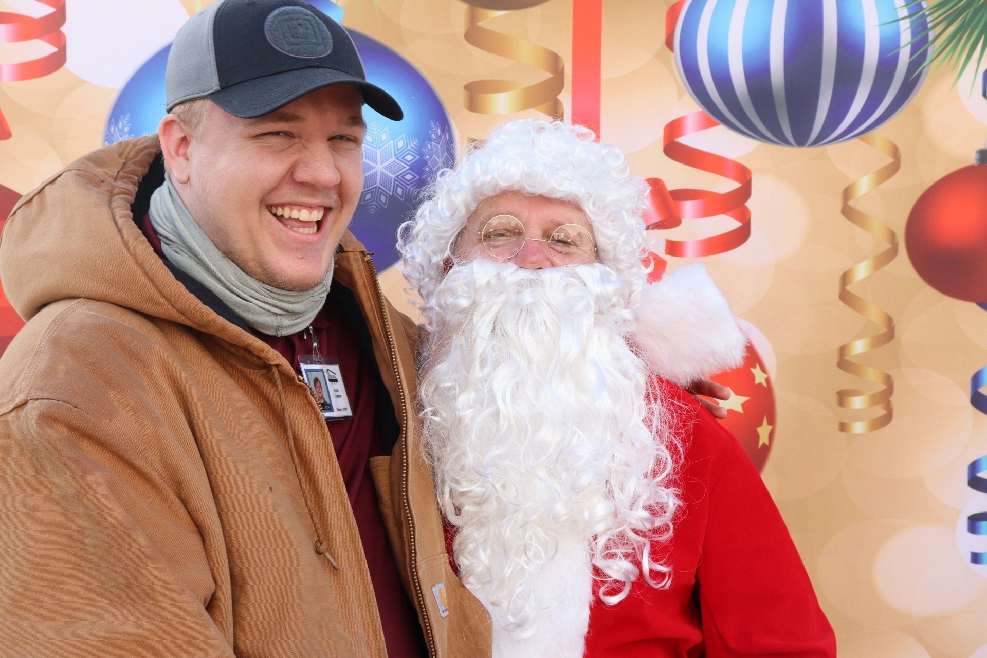 Man in workwear and Santa Claus smile at the camera, holiday decorations background.