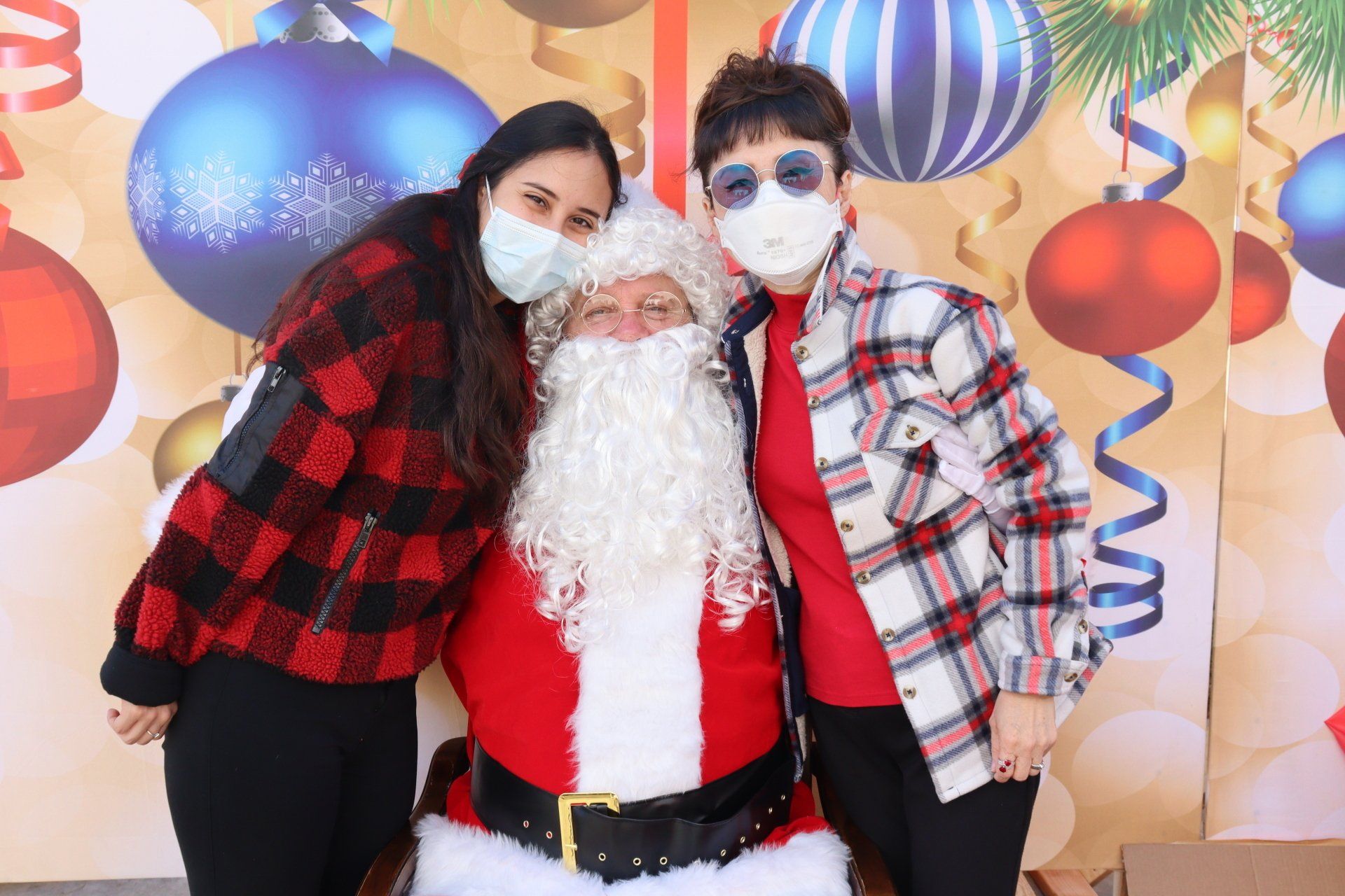 Two women in masks pose with Santa, against a Christmas backdrop.