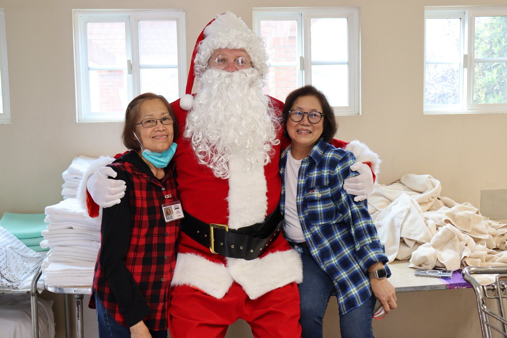 Santa Claus poses with two smiling women. Santa wears red suit; women wear casual attire. White setting.