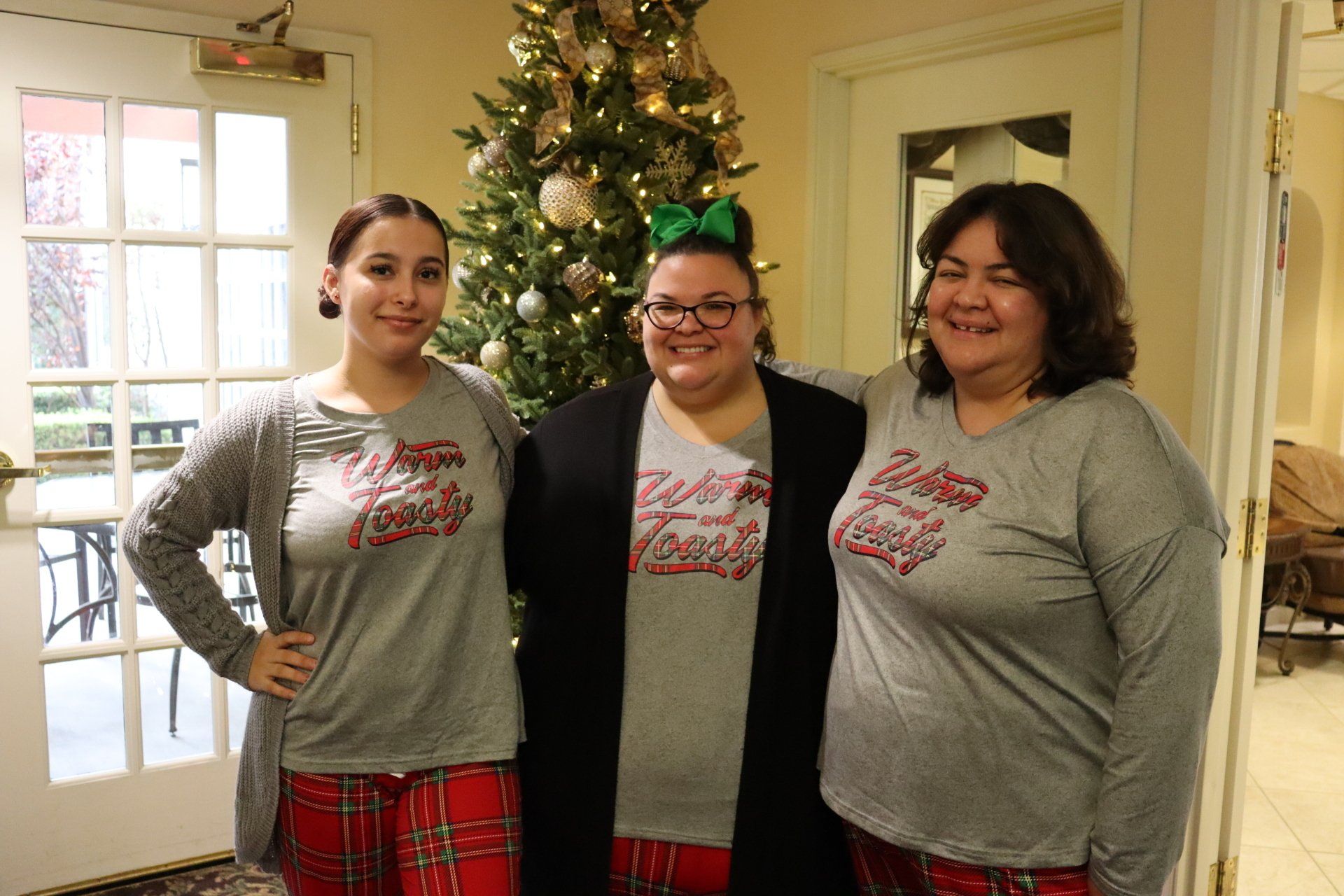 Three women in matching pajamas stand by a decorated Christmas tree indoors.