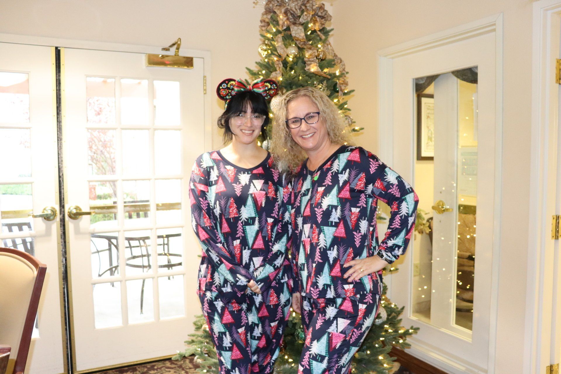 Two women in matching holiday pajamas pose by a Christmas tree, one wearing Mickey Mouse ears.