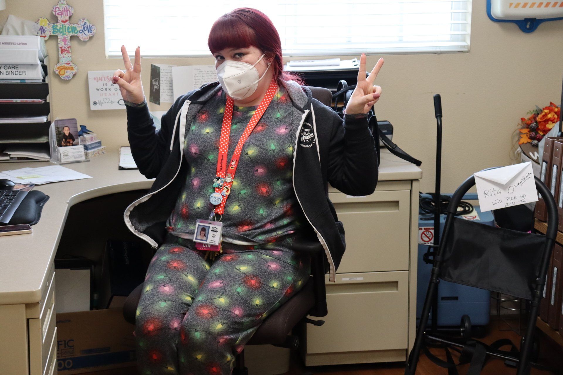 Person in pajamas and mask at desk, giving peace signs. Indoor office setting.