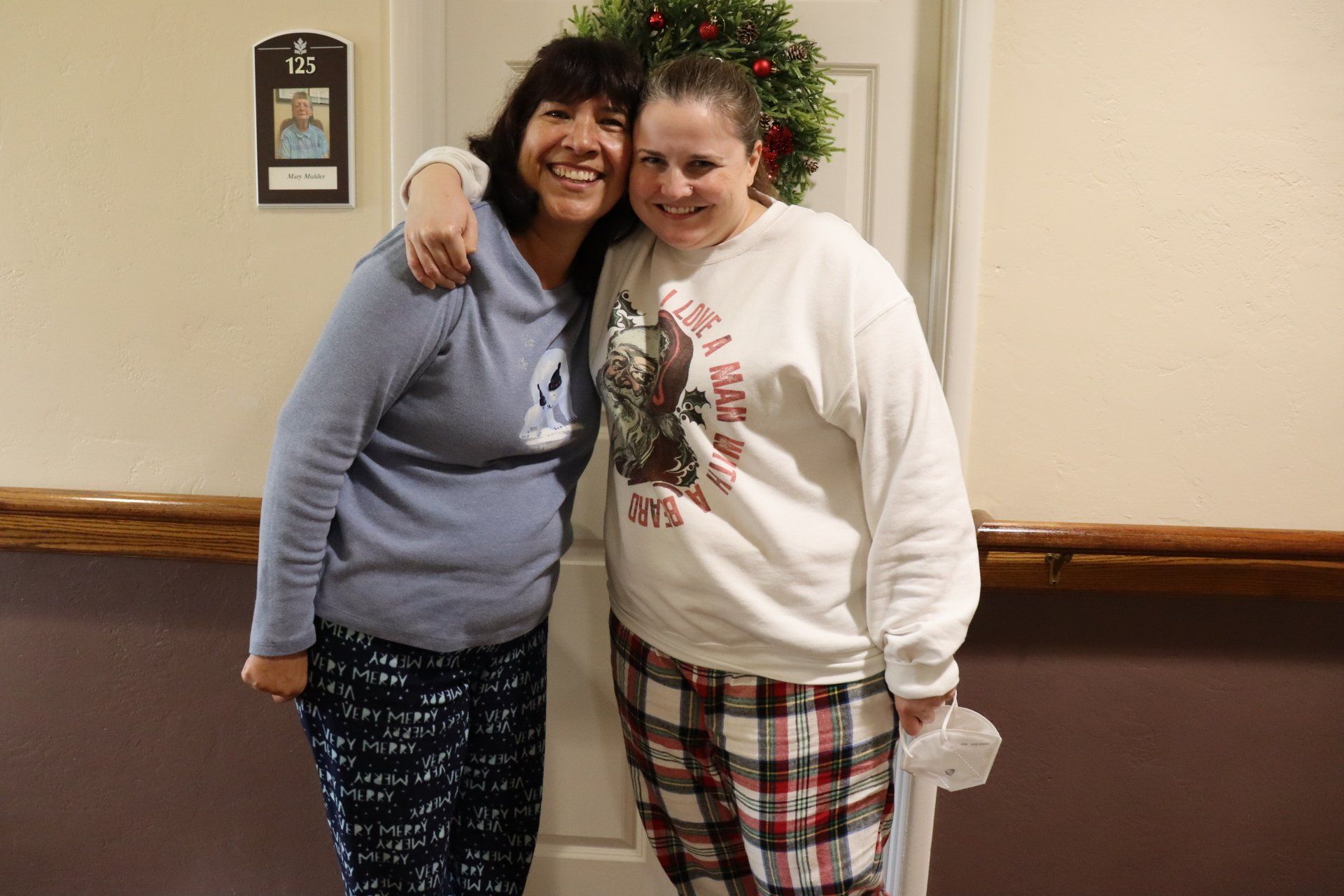 Two women in pajamas smile, embrace in front of a door decorated with a wreath.