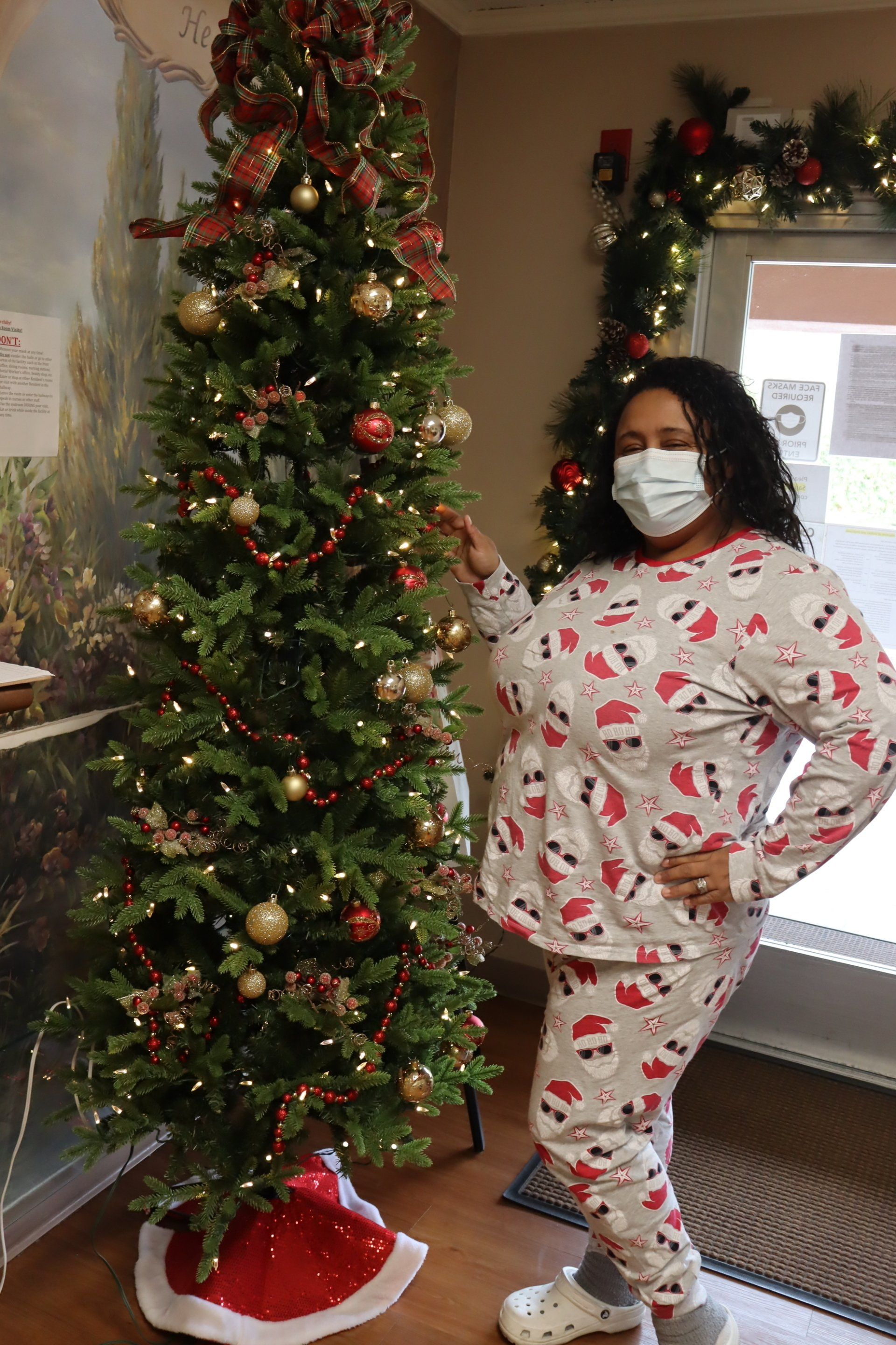 Woman in festive pajamas and mask poses next to a decorated Christmas tree.