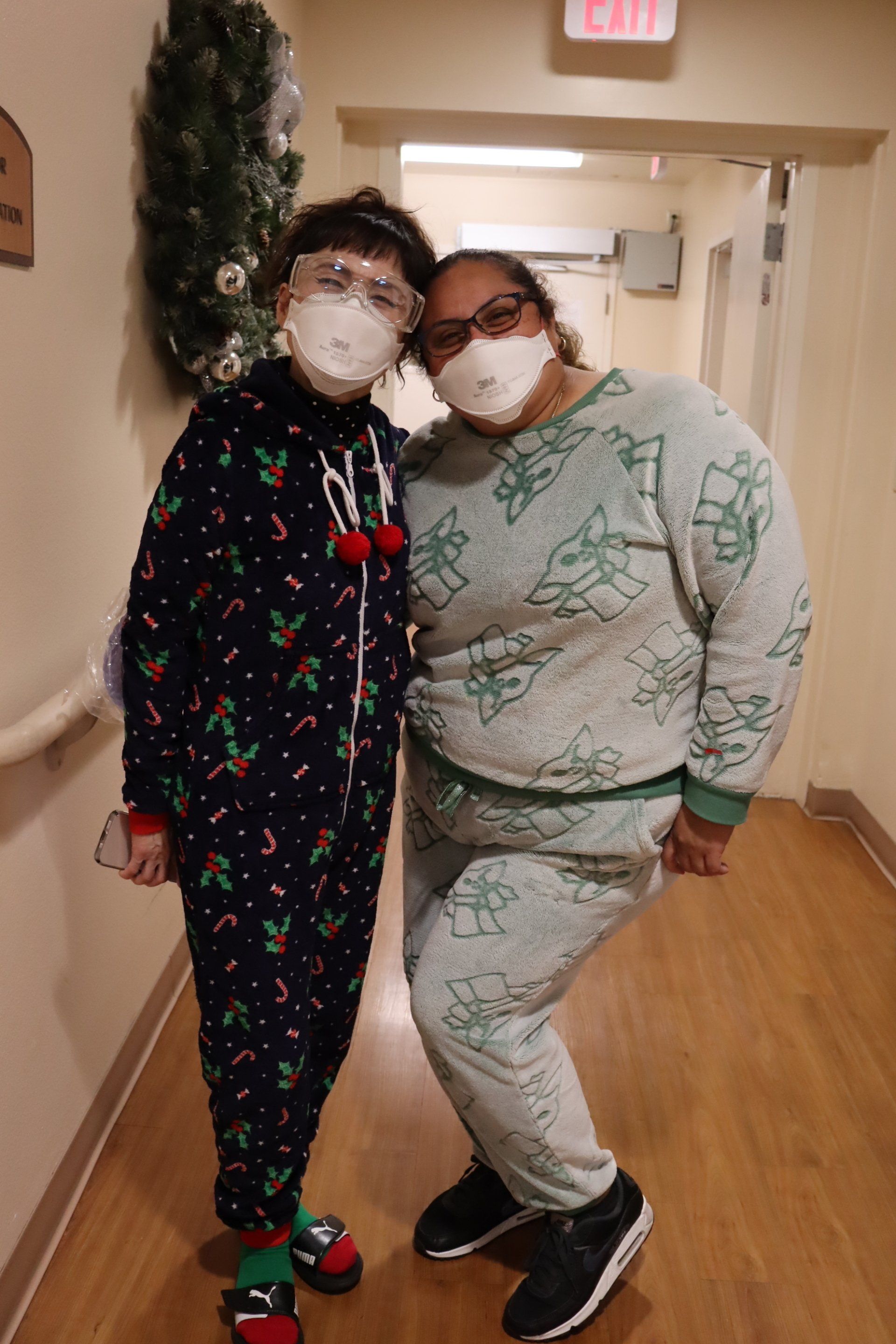Two people in pajamas pose in a hallway, wearing masks, one by a Christmas wreath.