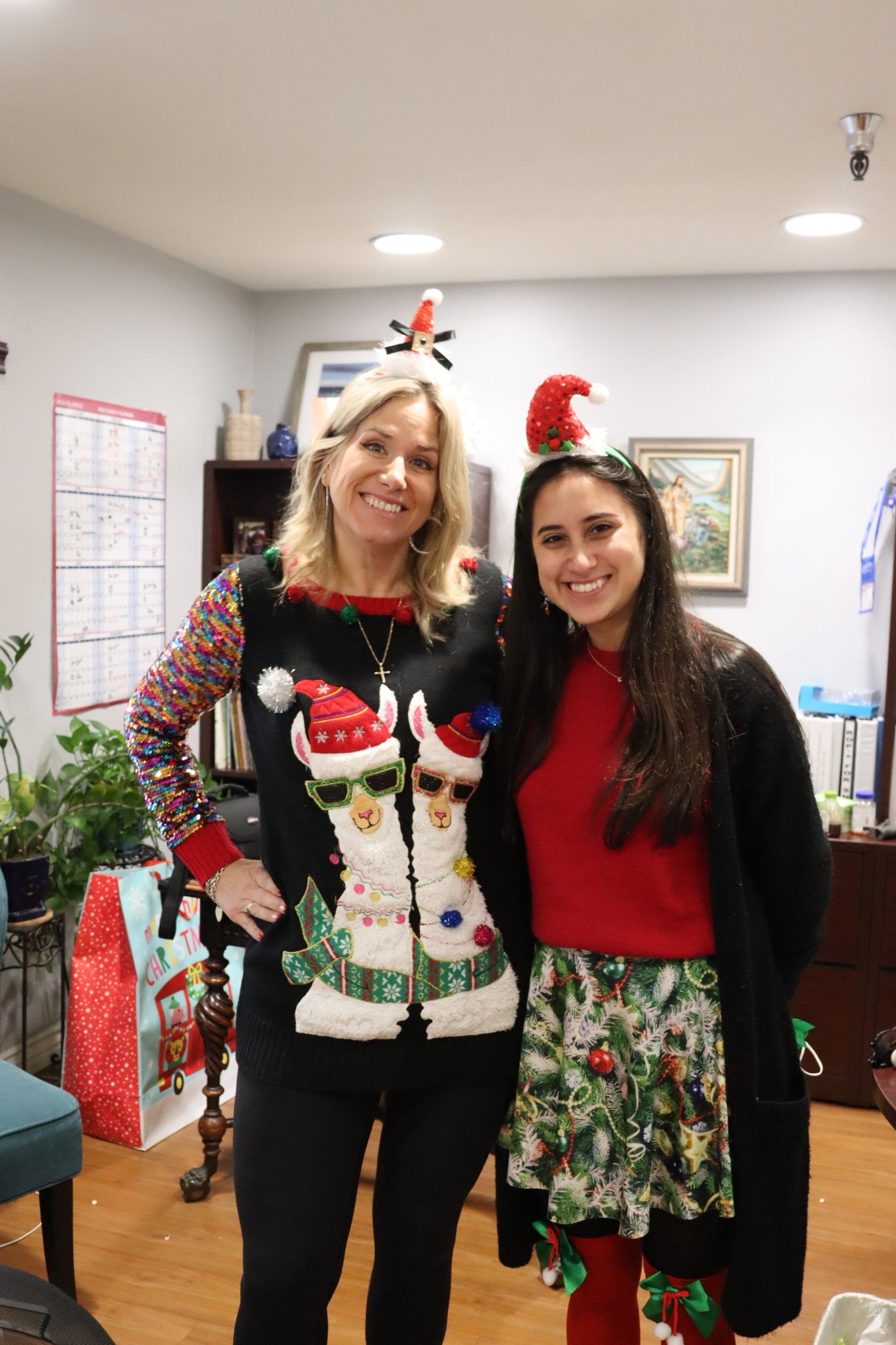 Two women in festive Christmas attire pose indoors; one in sweater with snowmen, other in red sweater and skirt.