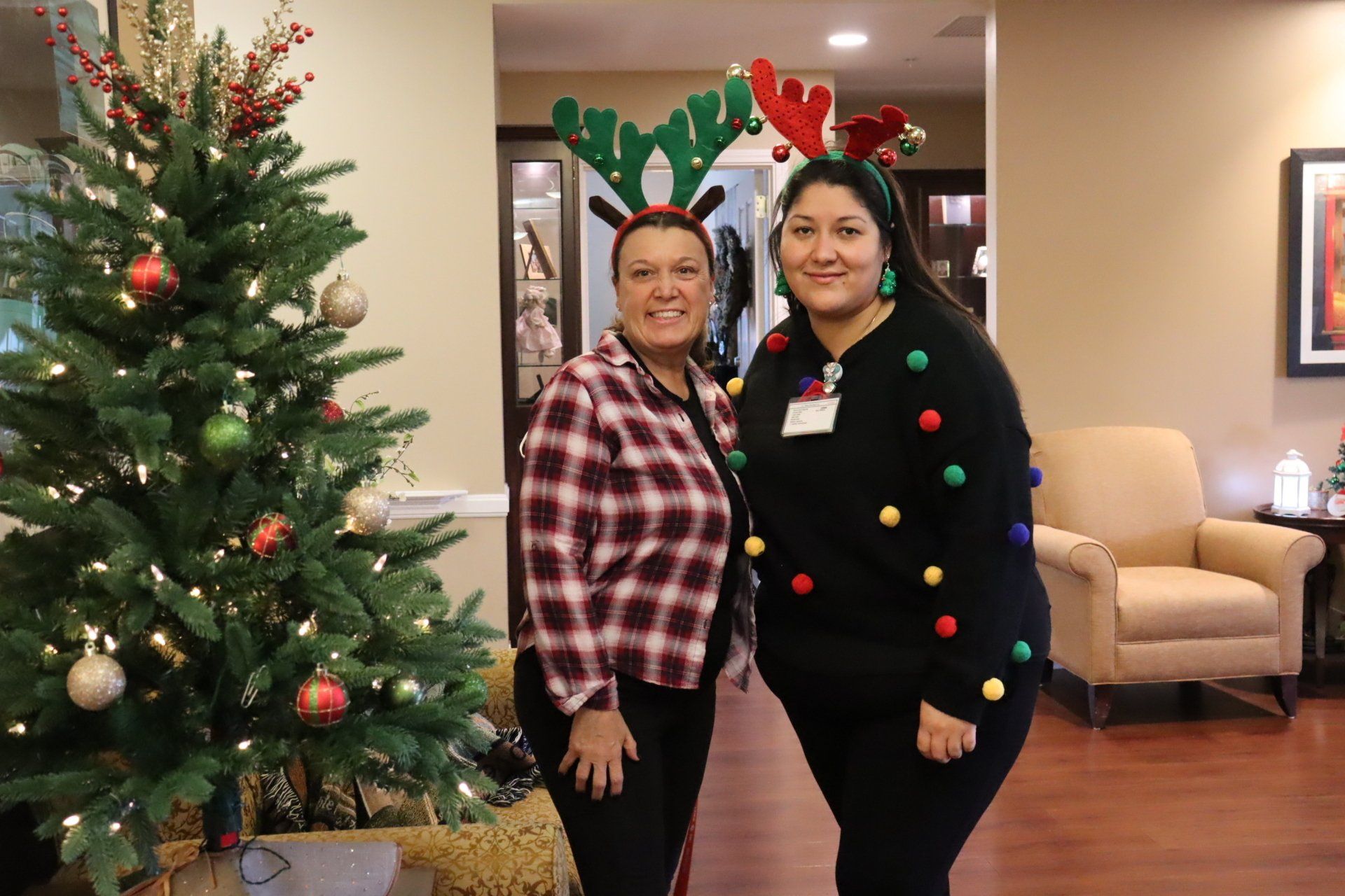 Two women wearing reindeer antlers pose by a Christmas tree indoors.