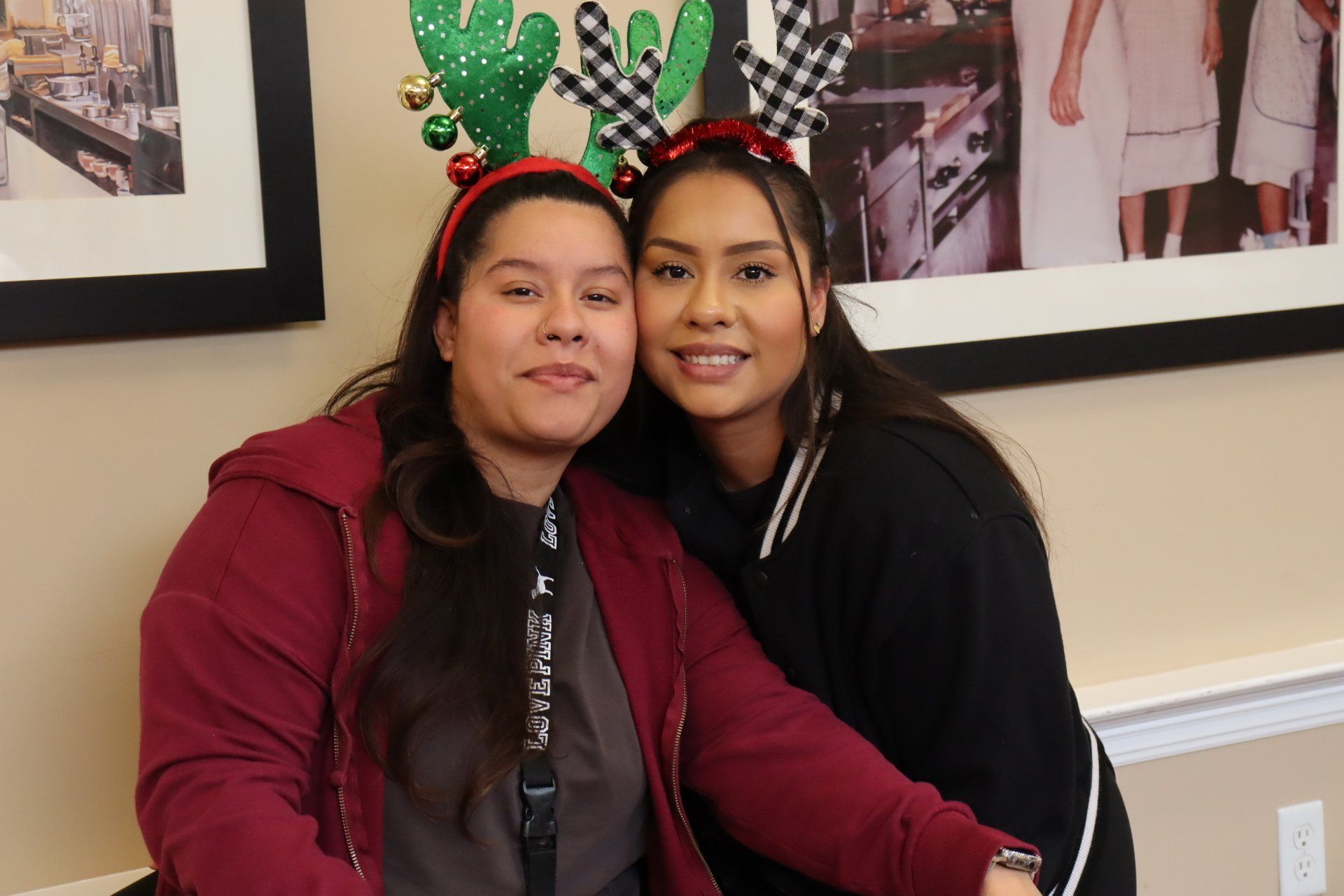 Two women smiling, wearing holiday headbands; indoors, beige walls.