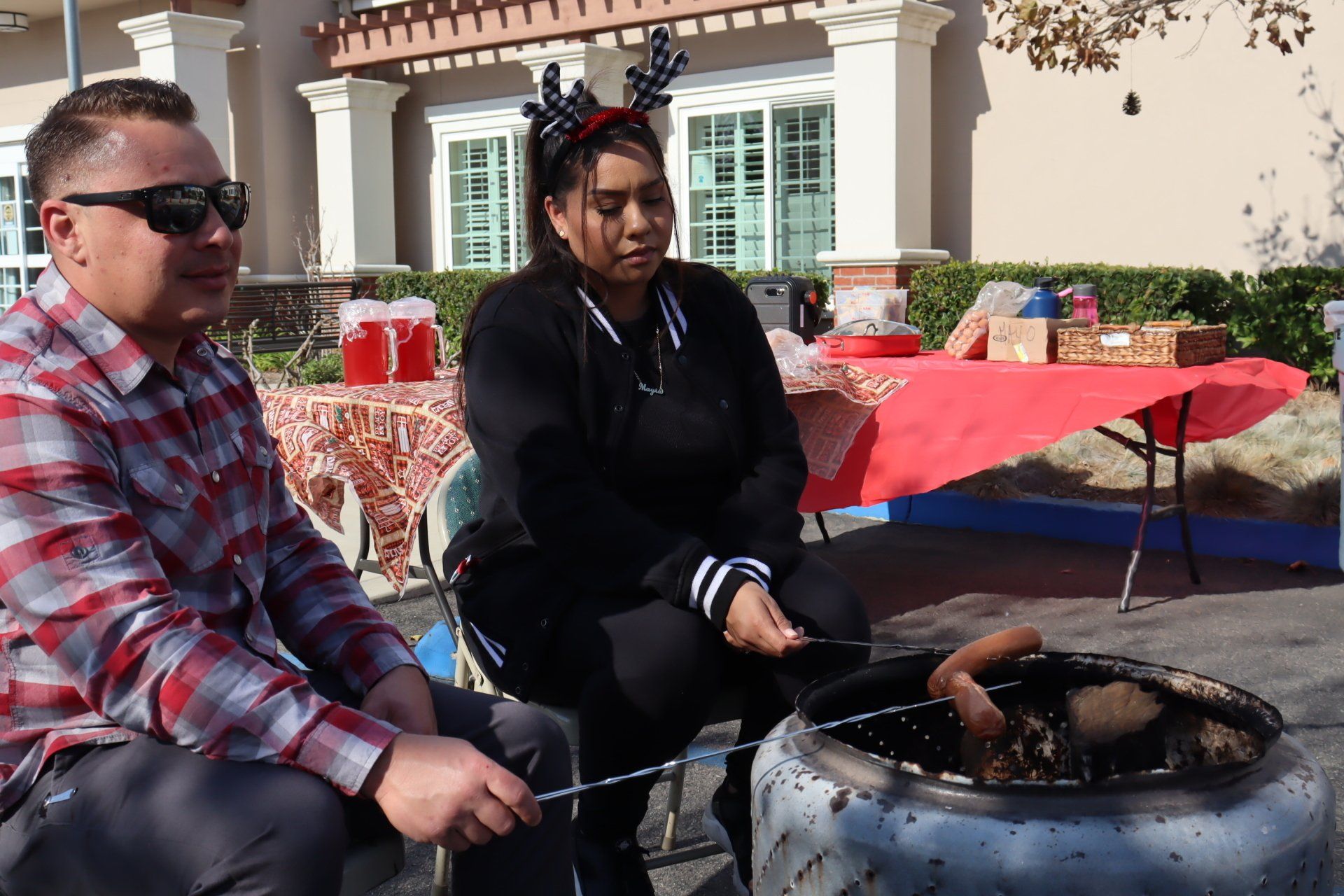 Man and woman roast hot dogs over a fire outdoors, near a table with gifts.