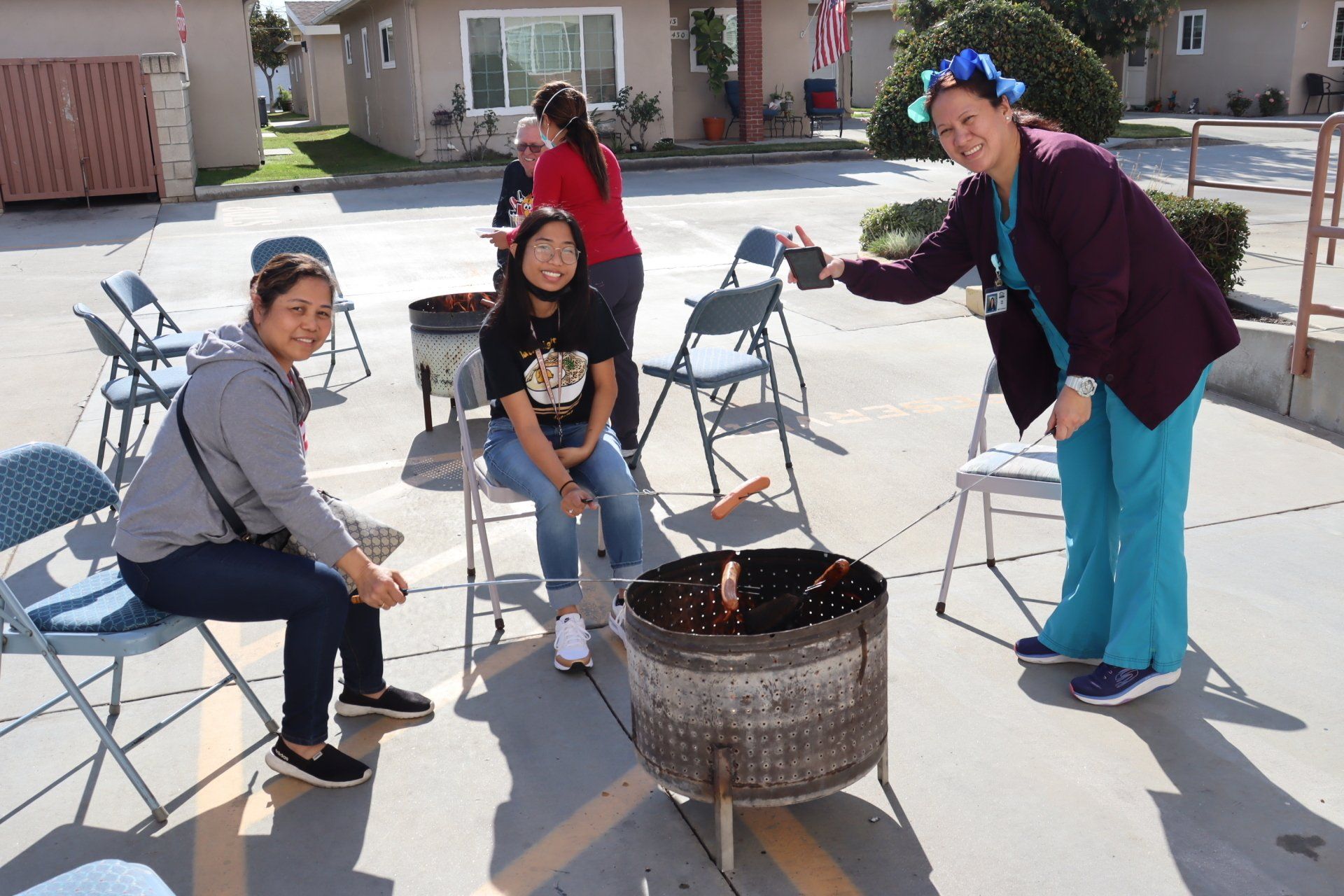 People outdoors around a grill. Woman in scrubs offers a drink. Others sit in chairs.