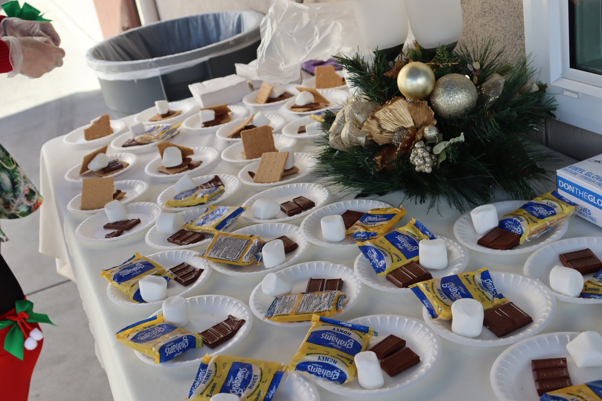 Table set with s'mores ingredients on plates, decorated with Christmas ornaments, outdoors.