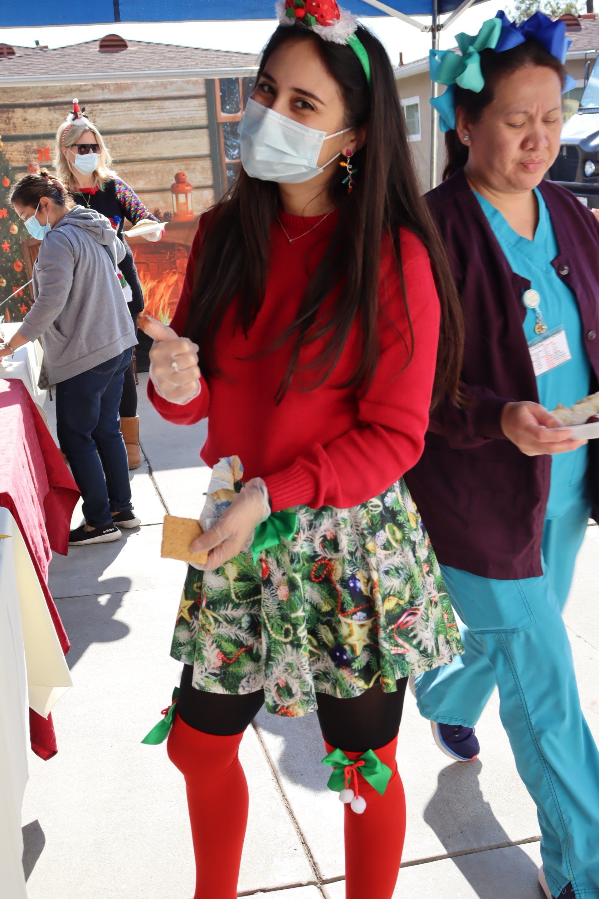 Woman in Christmas attire, mask on, gives thumbs up, standing outdoors with others.