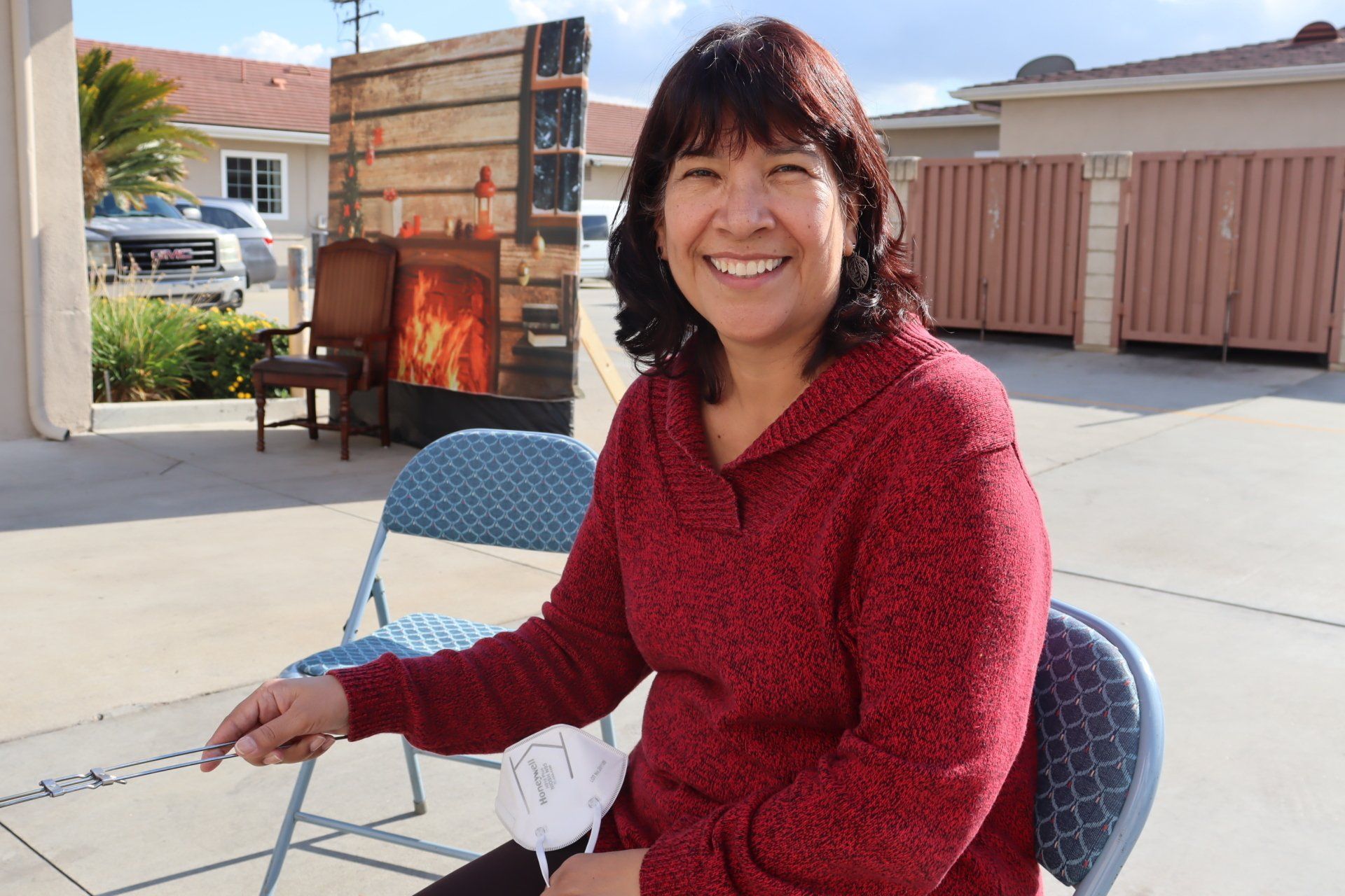Woman in red sweater smiles, seated outdoors. A painted fireplace and chair are behind her.