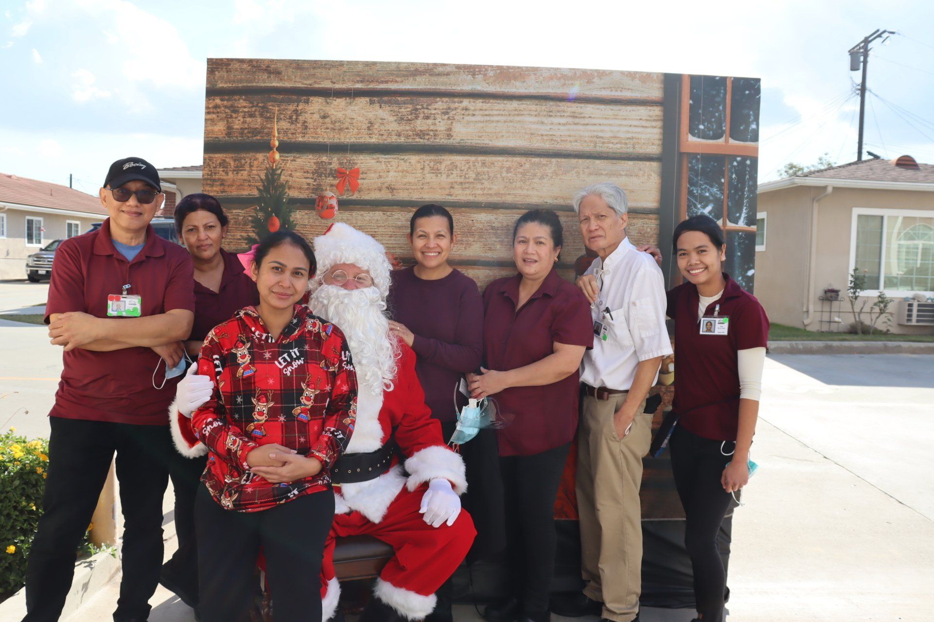 Santa with group of people in front of wooden backdrop. Many are wearing maroon shirts. Outdoors.