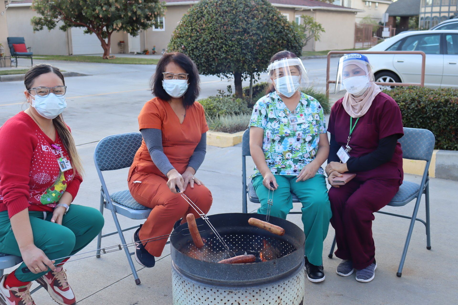 Four medical staff members wearing masks and scrubs grill hot dogs outdoors.
