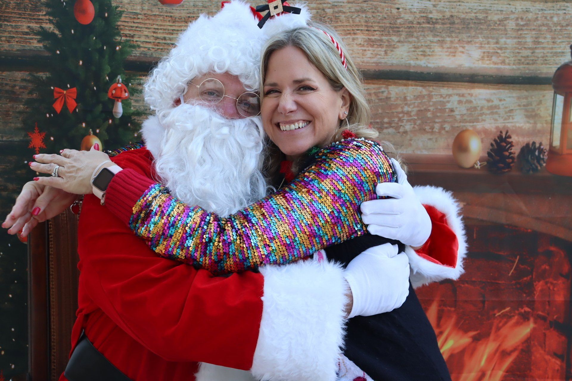Santa Claus hugging a smiling woman with a Christmas tree and fireplace backdrop.