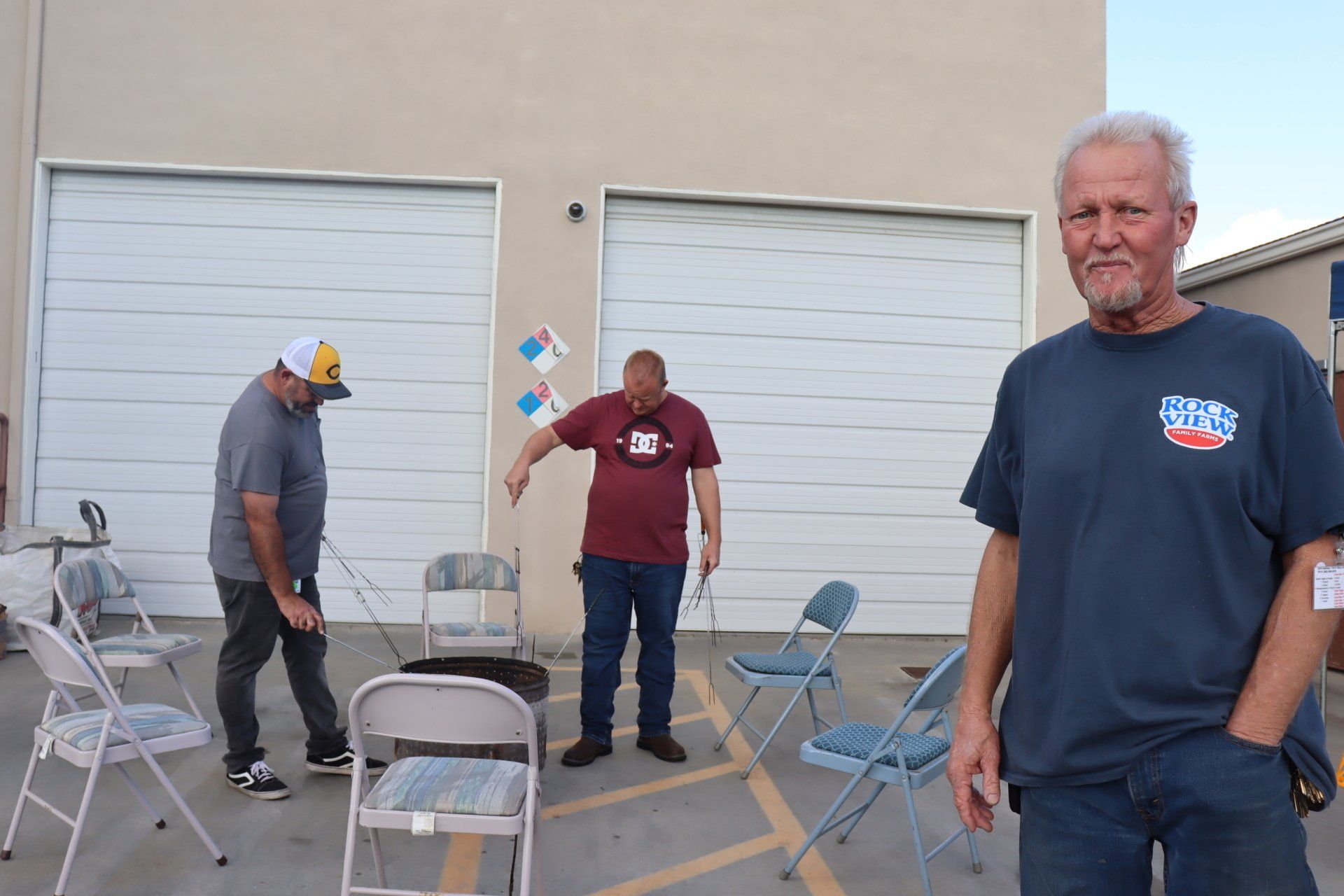 Three men standing outside, near chairs and garage doors. One man is in the foreground, looking at the camera.