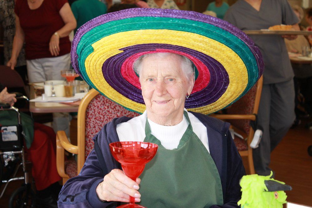 An elderly women holding a drink and wearing  sombrero.