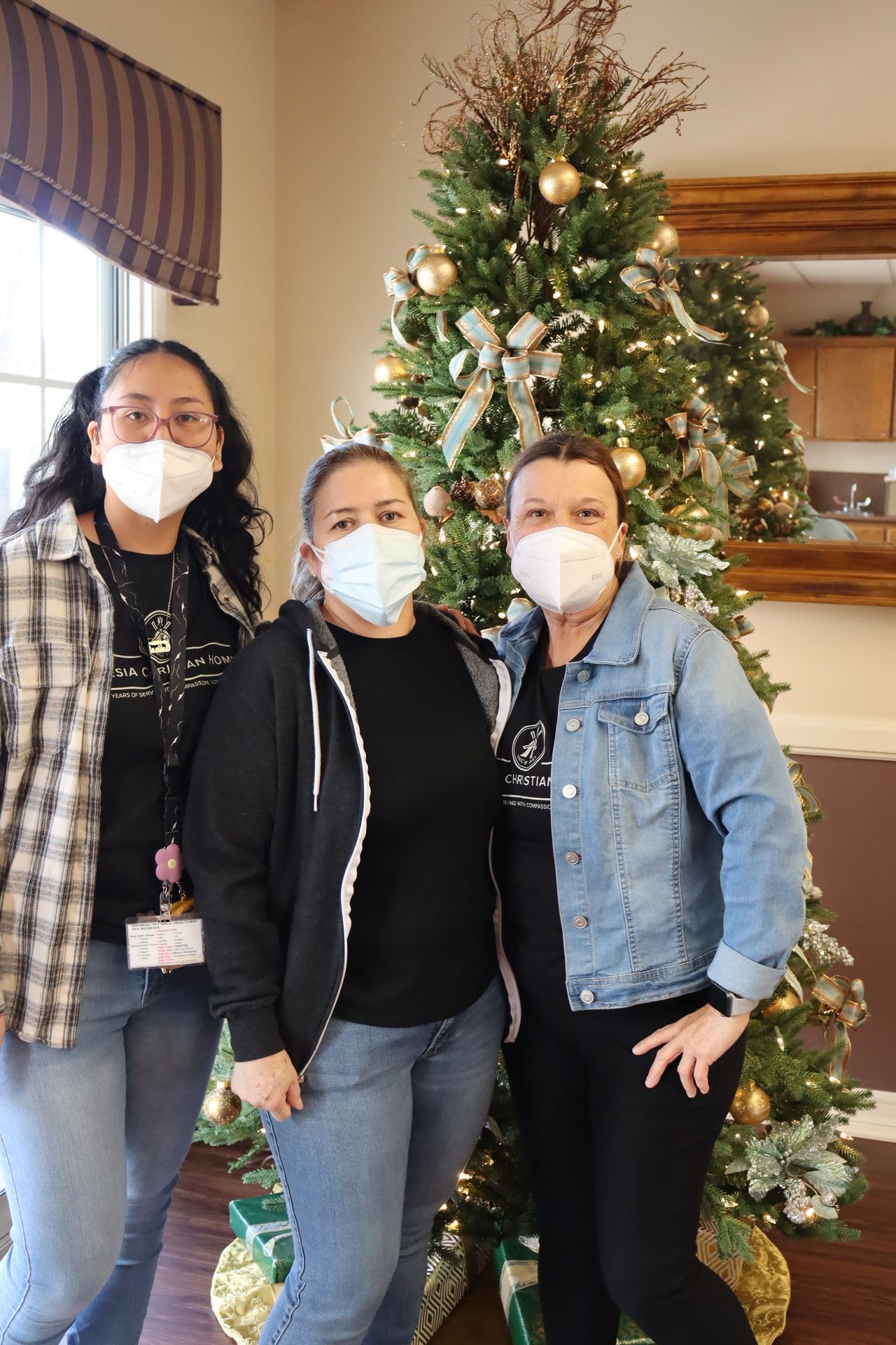 Three women wearing masks pose in front of a decorated Christmas tree.