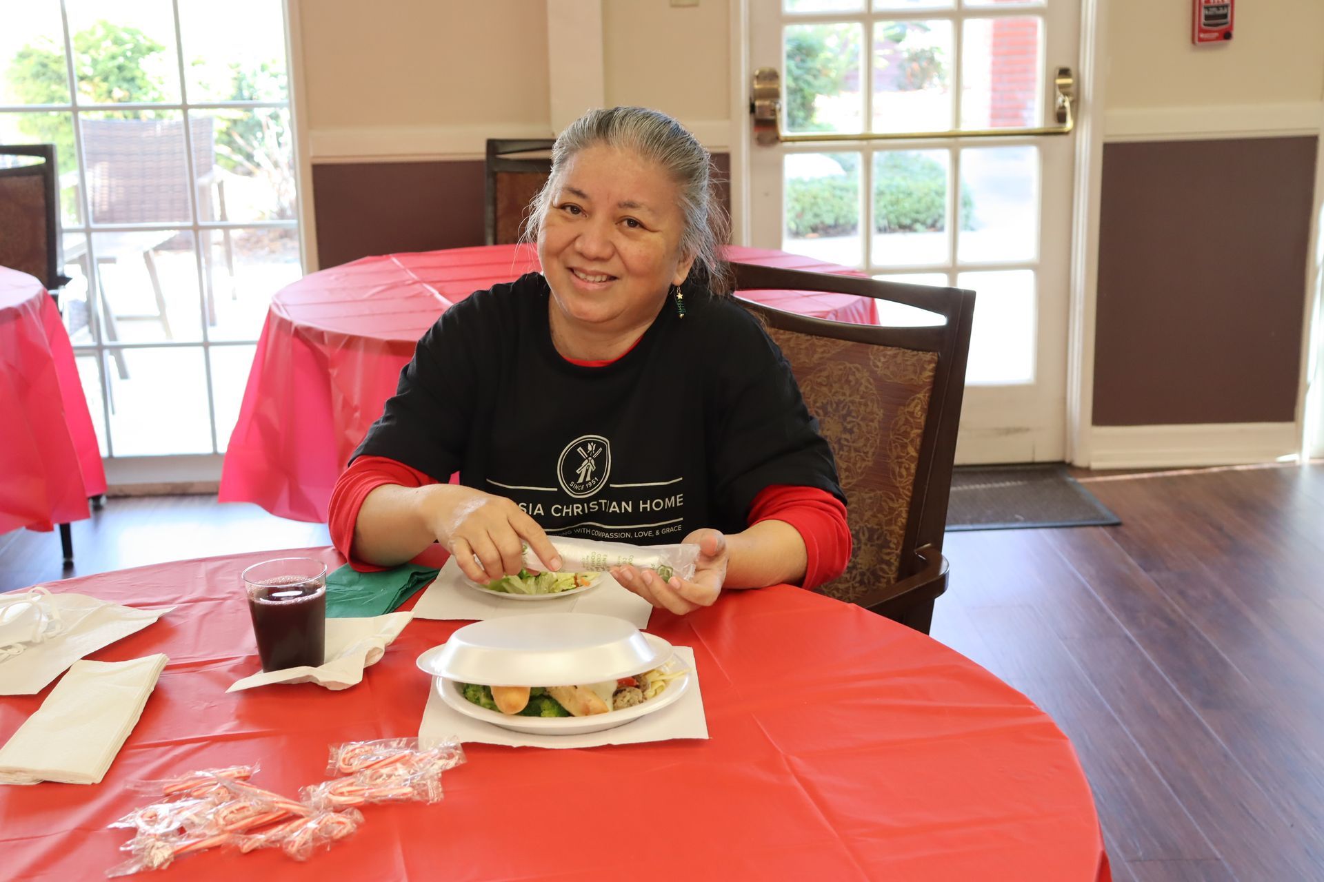 Woman smiling at a table with food, wearing black shirt, red tablecloth, indoor setting.