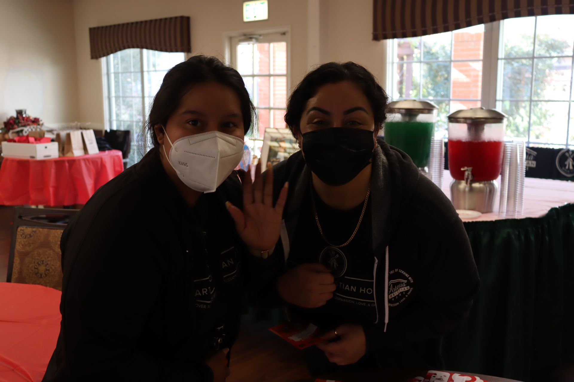 Two women in black shirts and masks wave, near beverage dispensers.