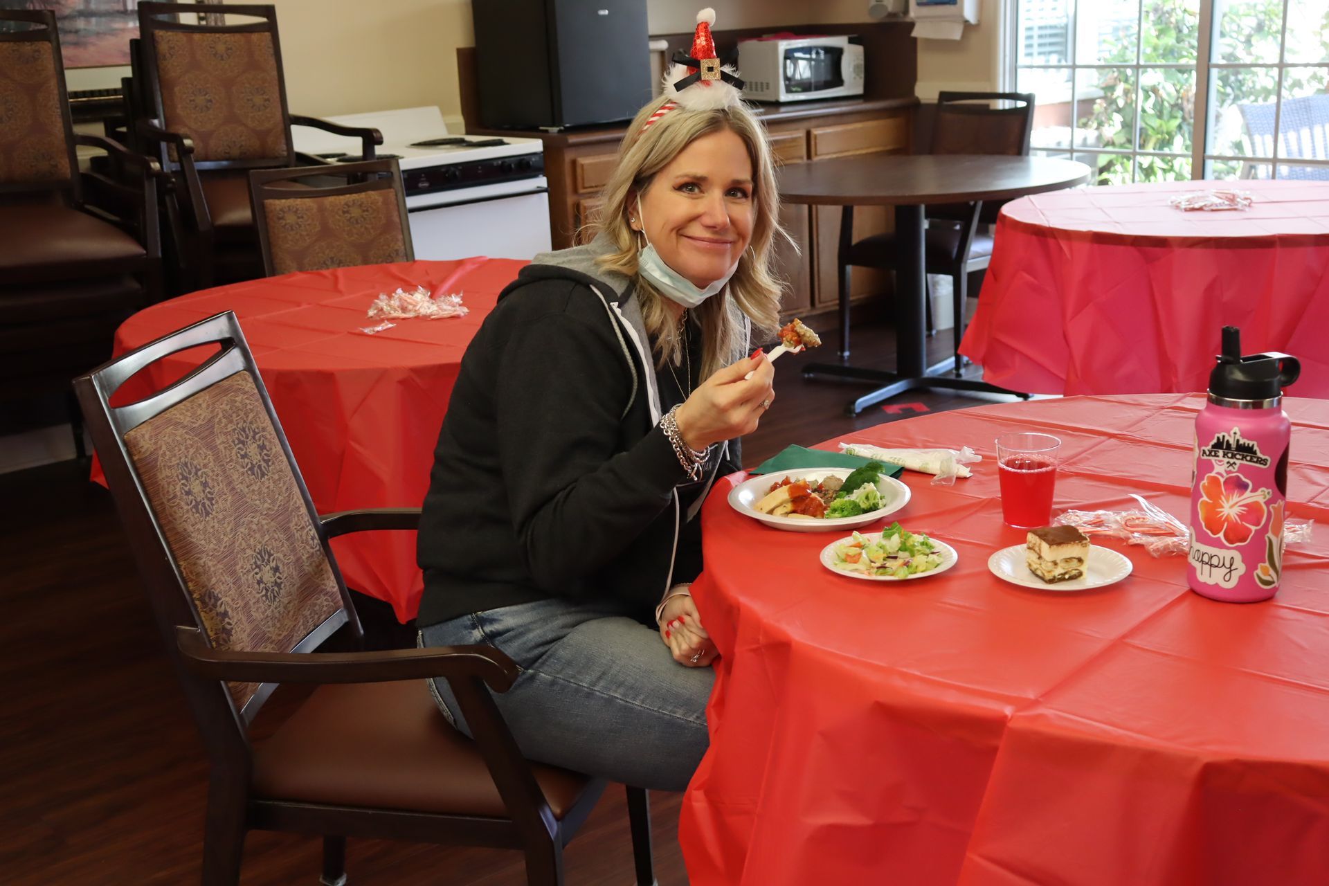 Woman at a red-covered table, eating. She wears a mask, black jacket, jeans, and a festive headband.