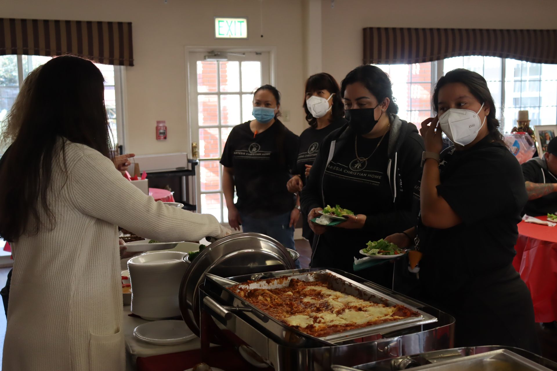 People at a buffet serving themselves lasagna. Some wear masks. Inside a room with a door.