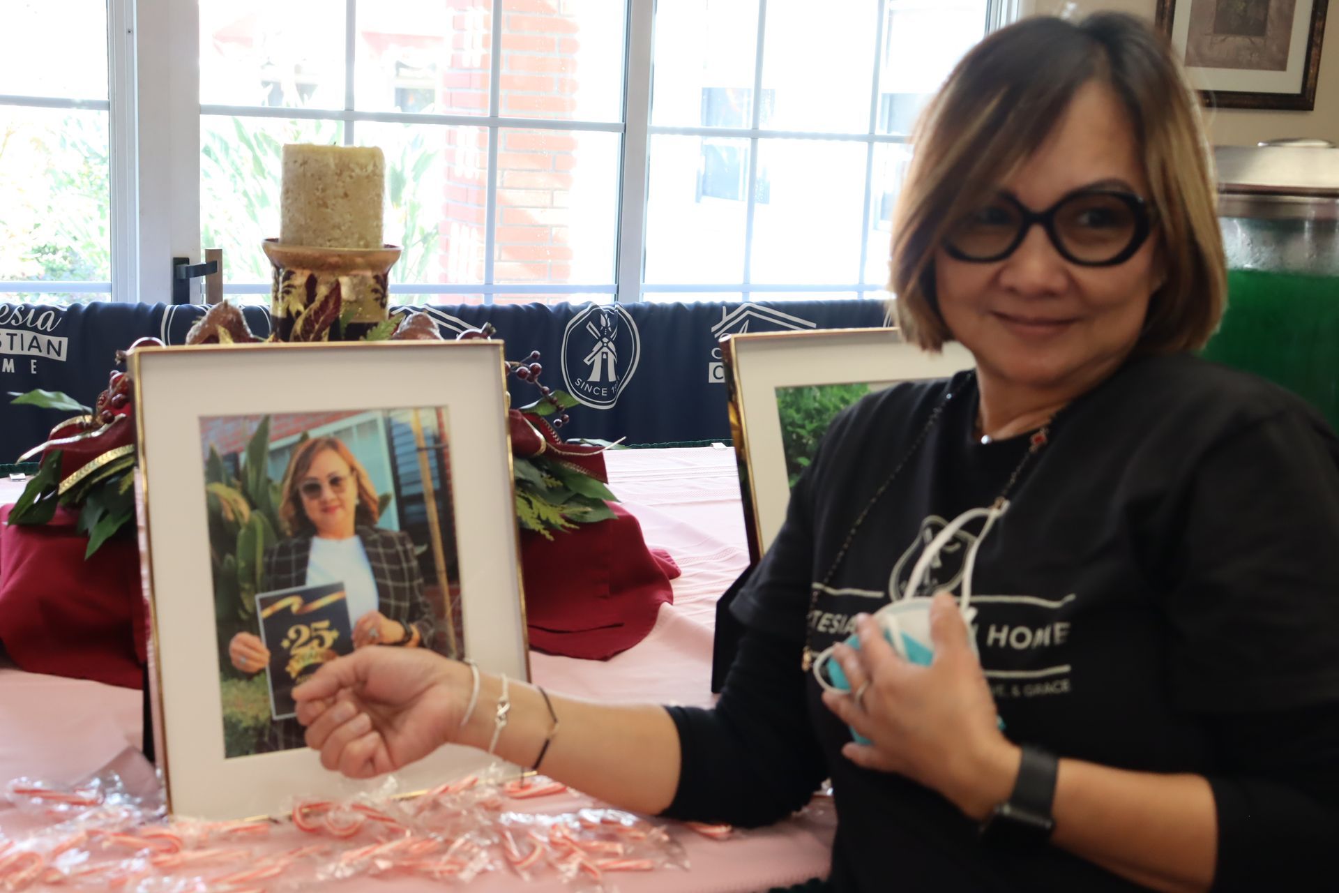 Woman with glasses, holding an object, smiles next to framed photos.  Candle and decorations on table.
