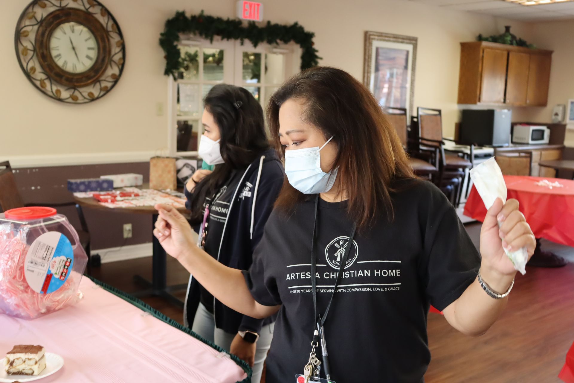 Two women in masks at a party. One points at a jar, the other stands nearby. Pink and red decorations.