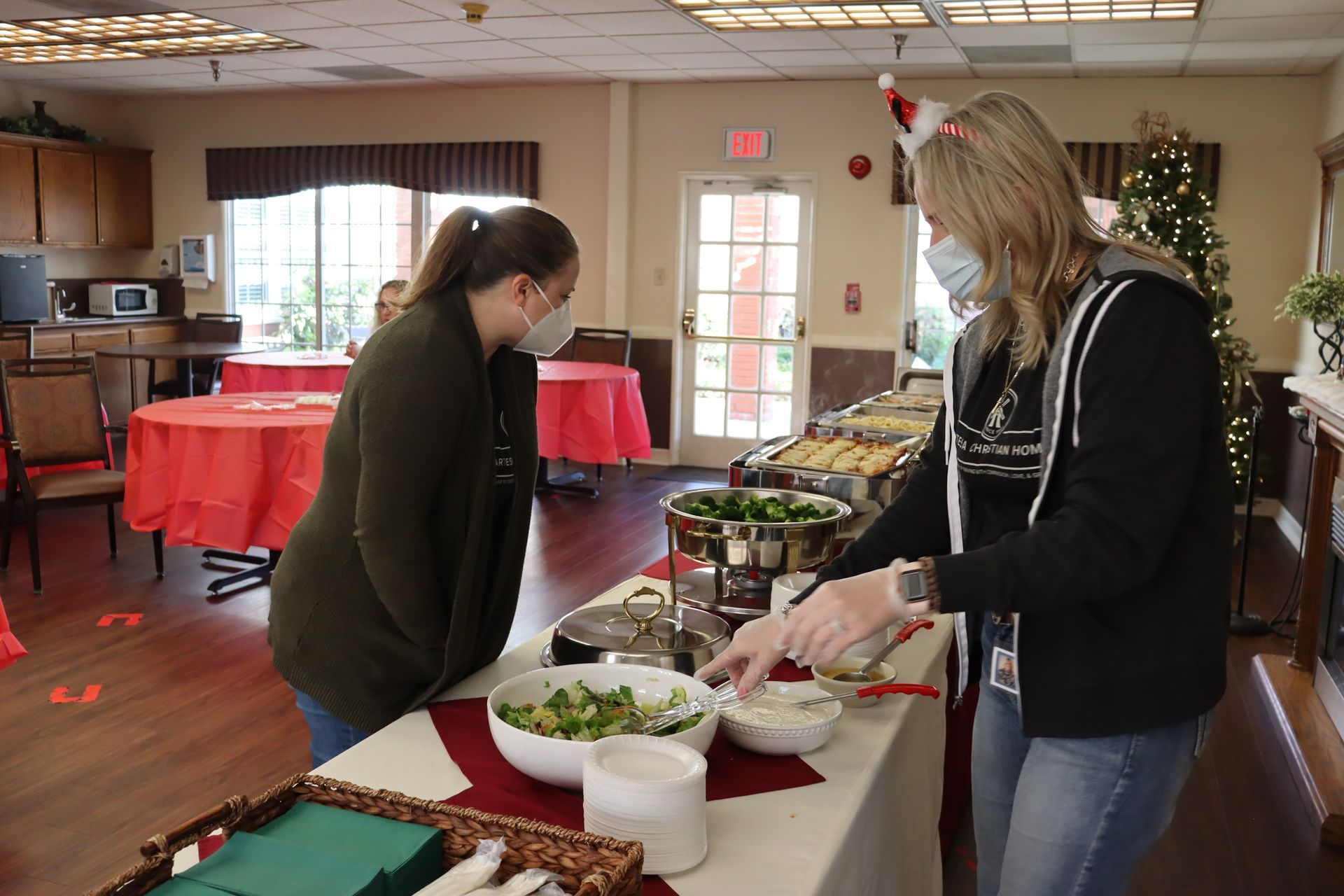Two women at a buffet, one serving salad. Both wear masks. Setting is a decorated dining room.
