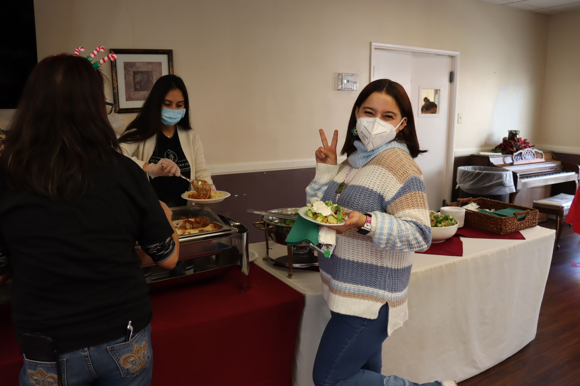 People at a buffet. Woman in a mask makes a peace sign, holding a plate. Others serve food, room is decorated.