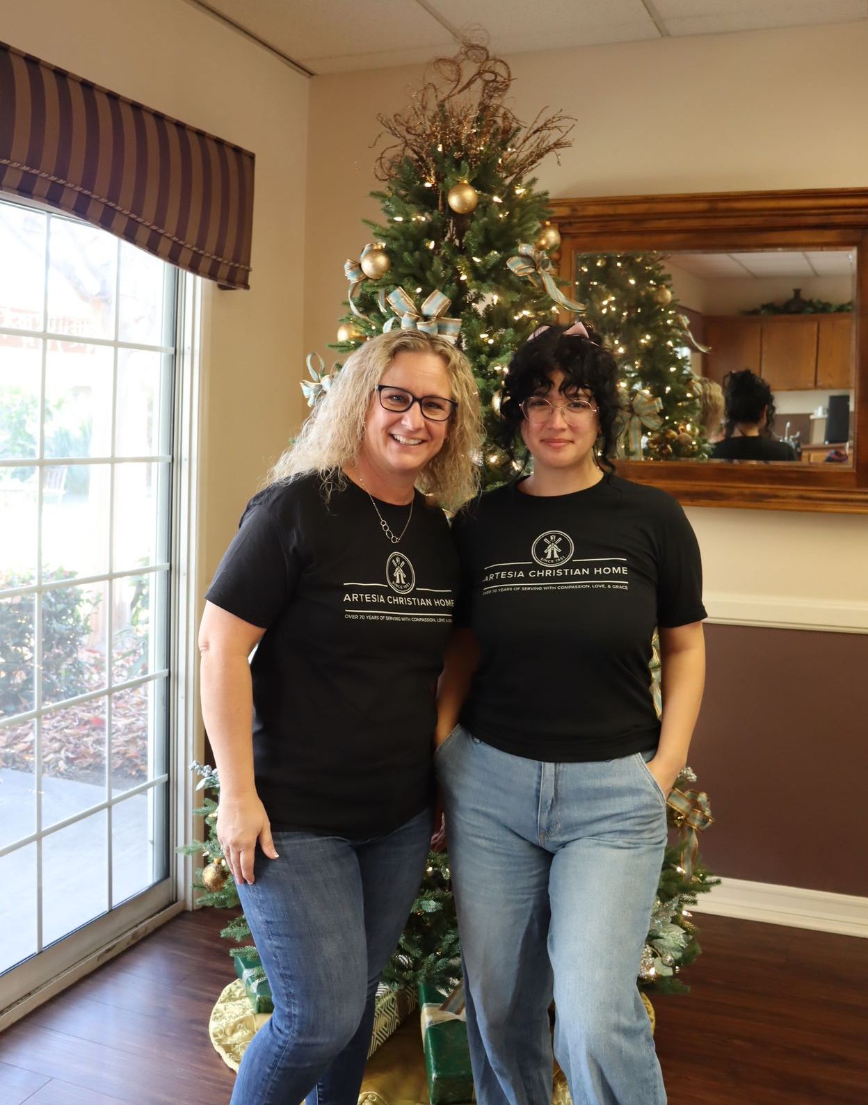 Two women in black shirts and jeans pose by a decorated Christmas tree and window.