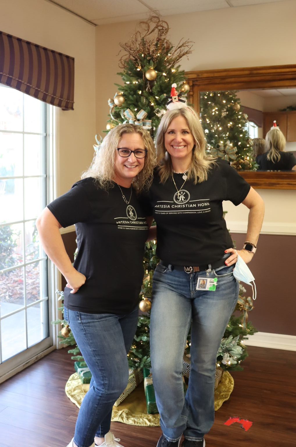 Two women in black shirts and jeans pose by a Christmas tree, inside a room with a mirror and window.