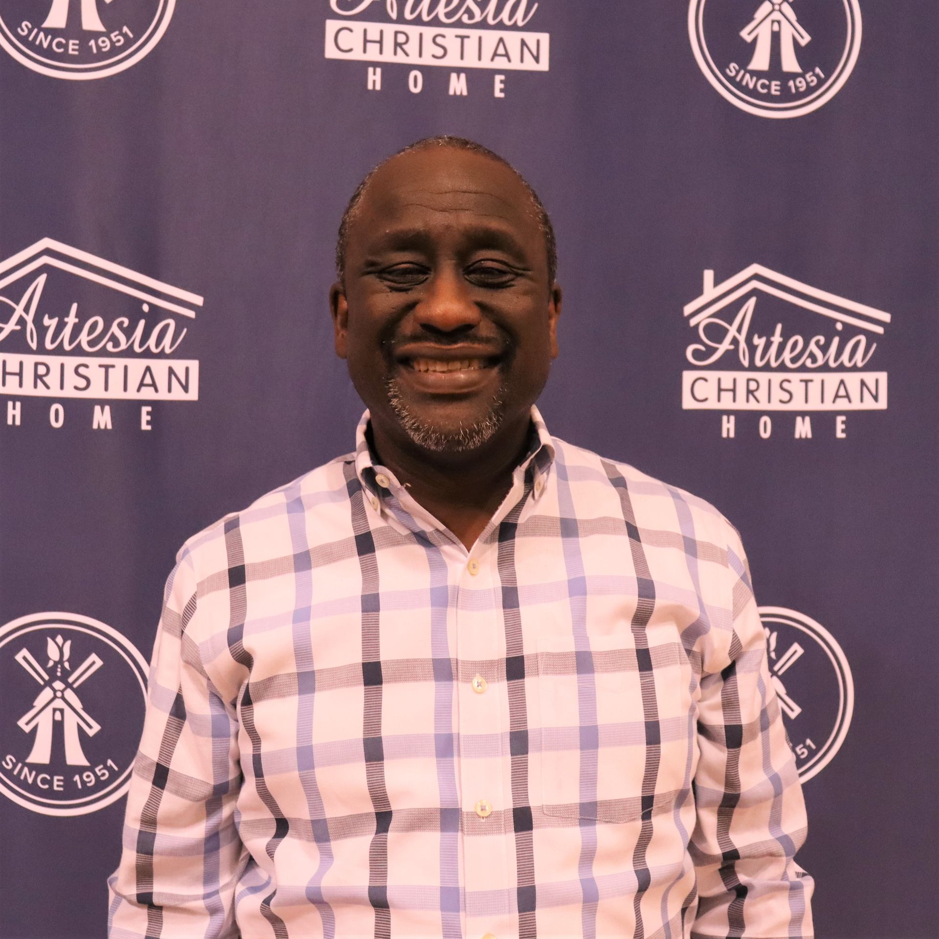 Smiling man with a salt-and-pepper beard, wearing a plaid shirt, in front of a blue backdrop with logos.