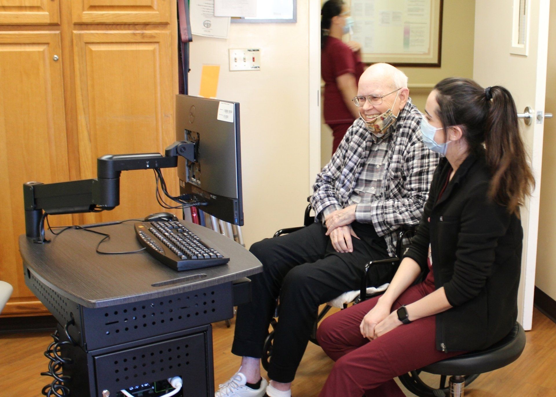 A healthcare worker helps an older man at a computer; both wearing masks.