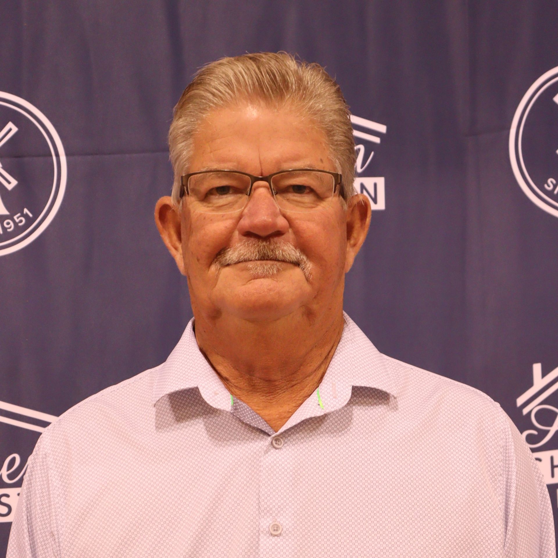 Senior man with glasses and mustache, light pink shirt, in front of a blue backdrop with logos.
